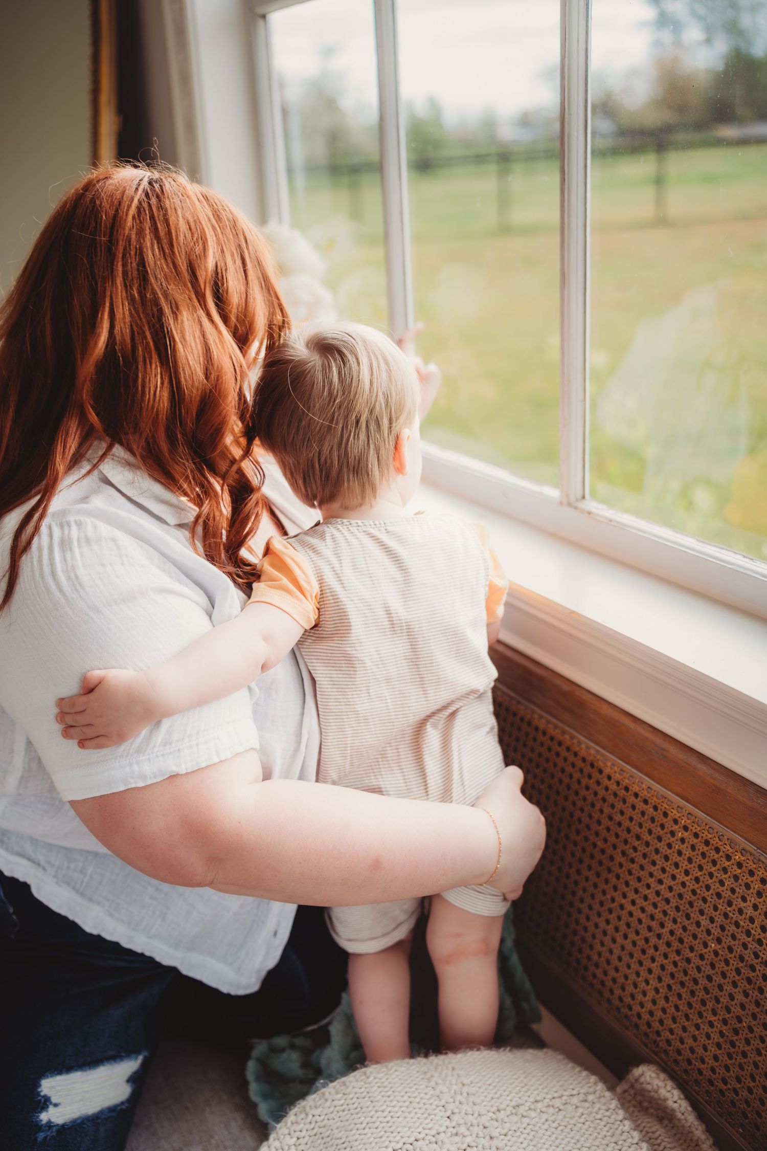 A red-haired woman holds a toddler while they look out a large window together at a green outdoor landscape.