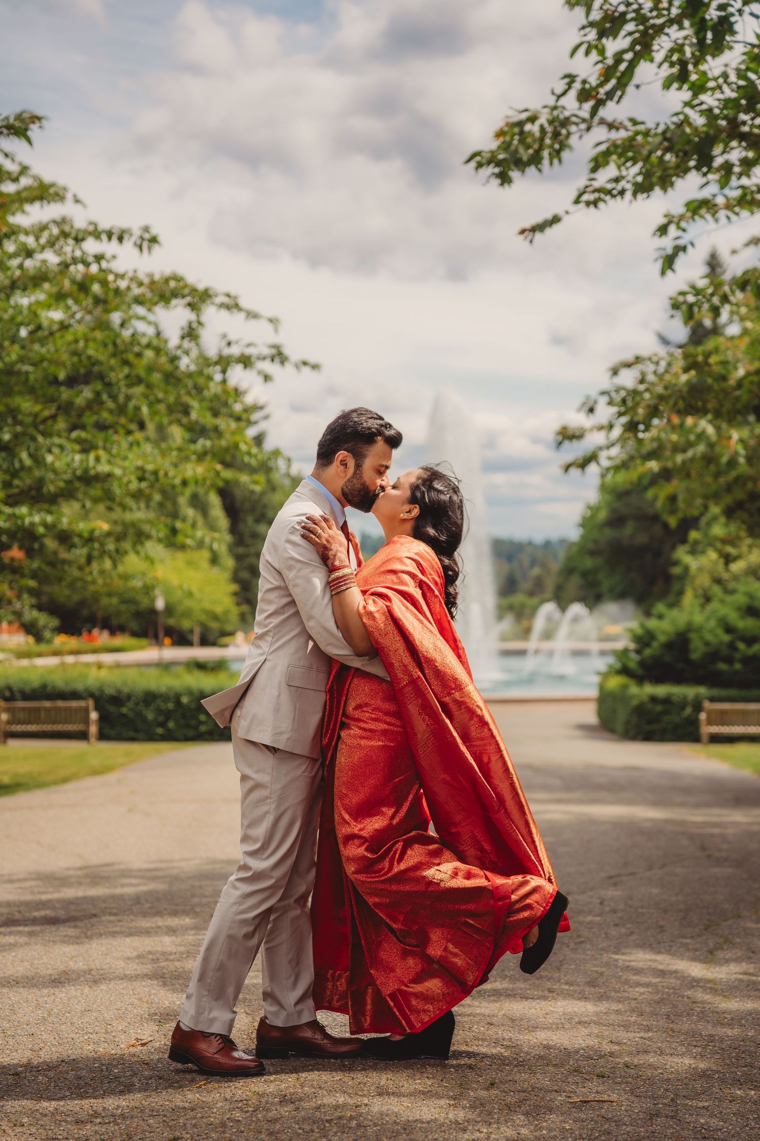 Couple in traditional Indian wedding attire sharing a romantic kiss in a lush garden park with a fountain backdrop.