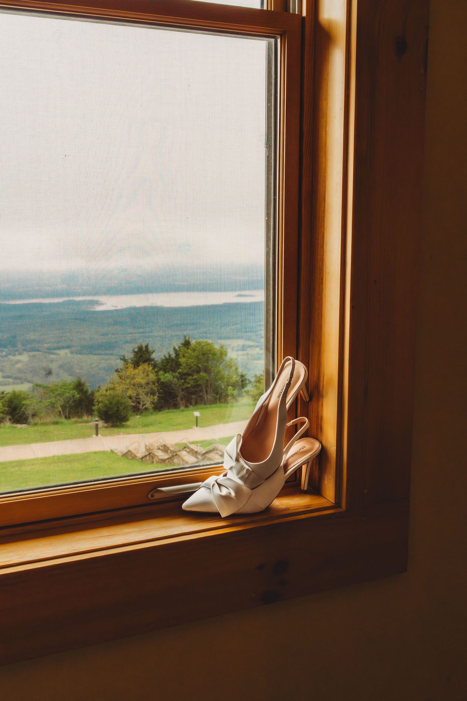 White bridal heels with bow detail resting on wooden windowsill overlooking scenic green hills and lake.