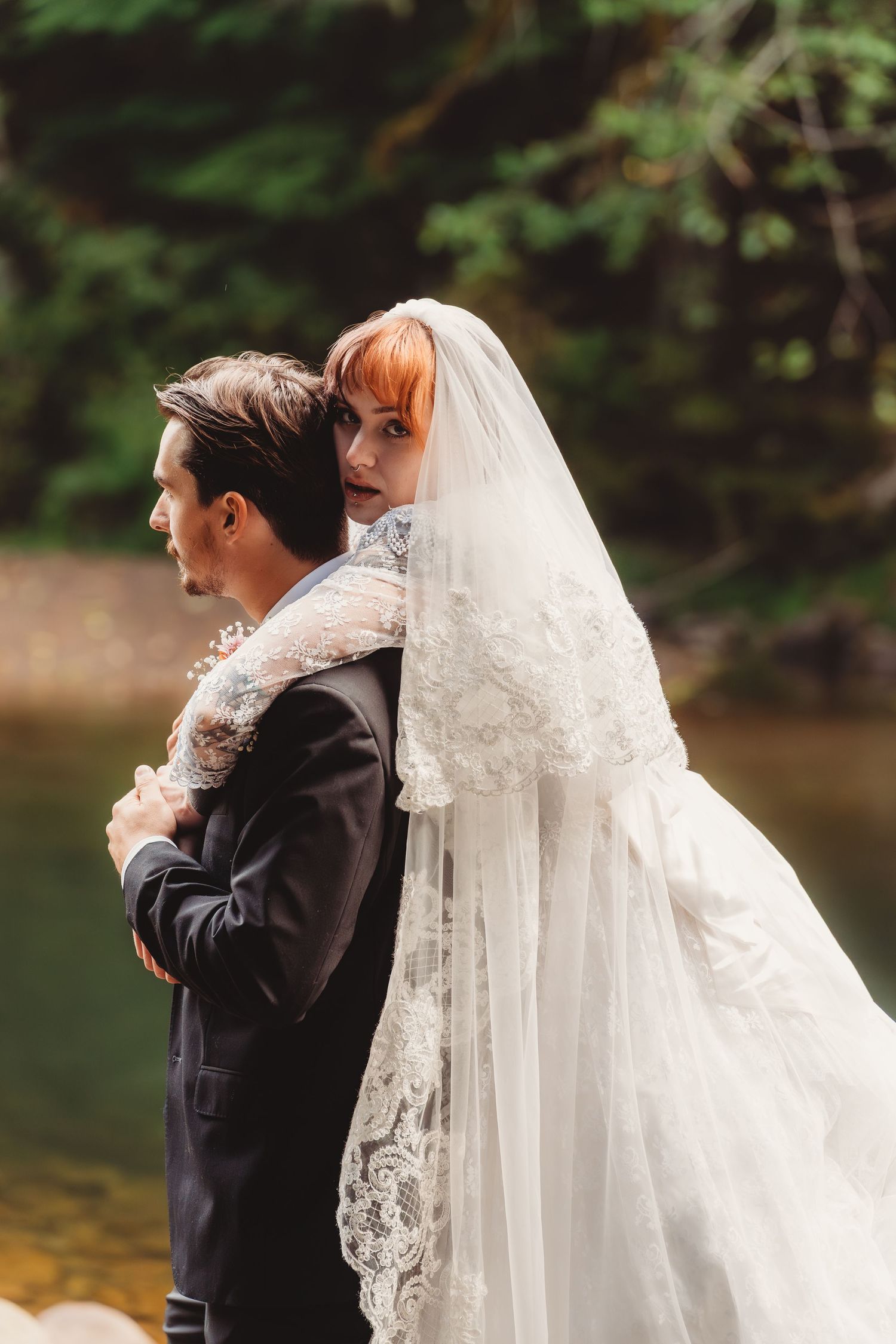 Bride in lace veil embraces groom in black suit outdoors near forest stream at wedding.