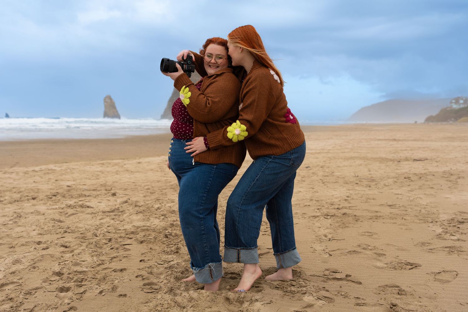 Three women in matching brown hoodies examine photos on a camera while standing barefoot on a sandy beach.