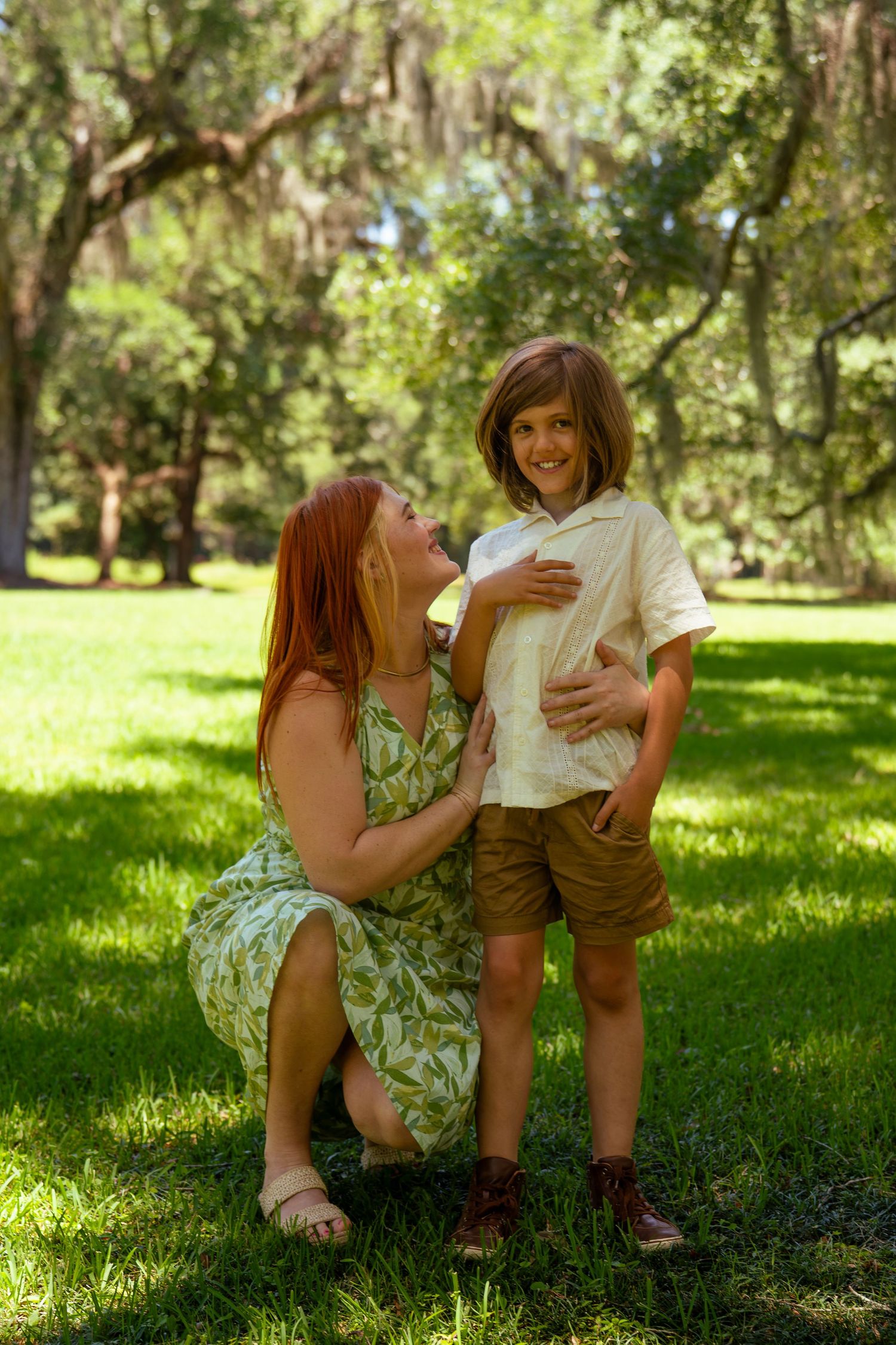 A redheaded woman kneels beside a smiling boy in a sunny green park with moss-draped oak trees.