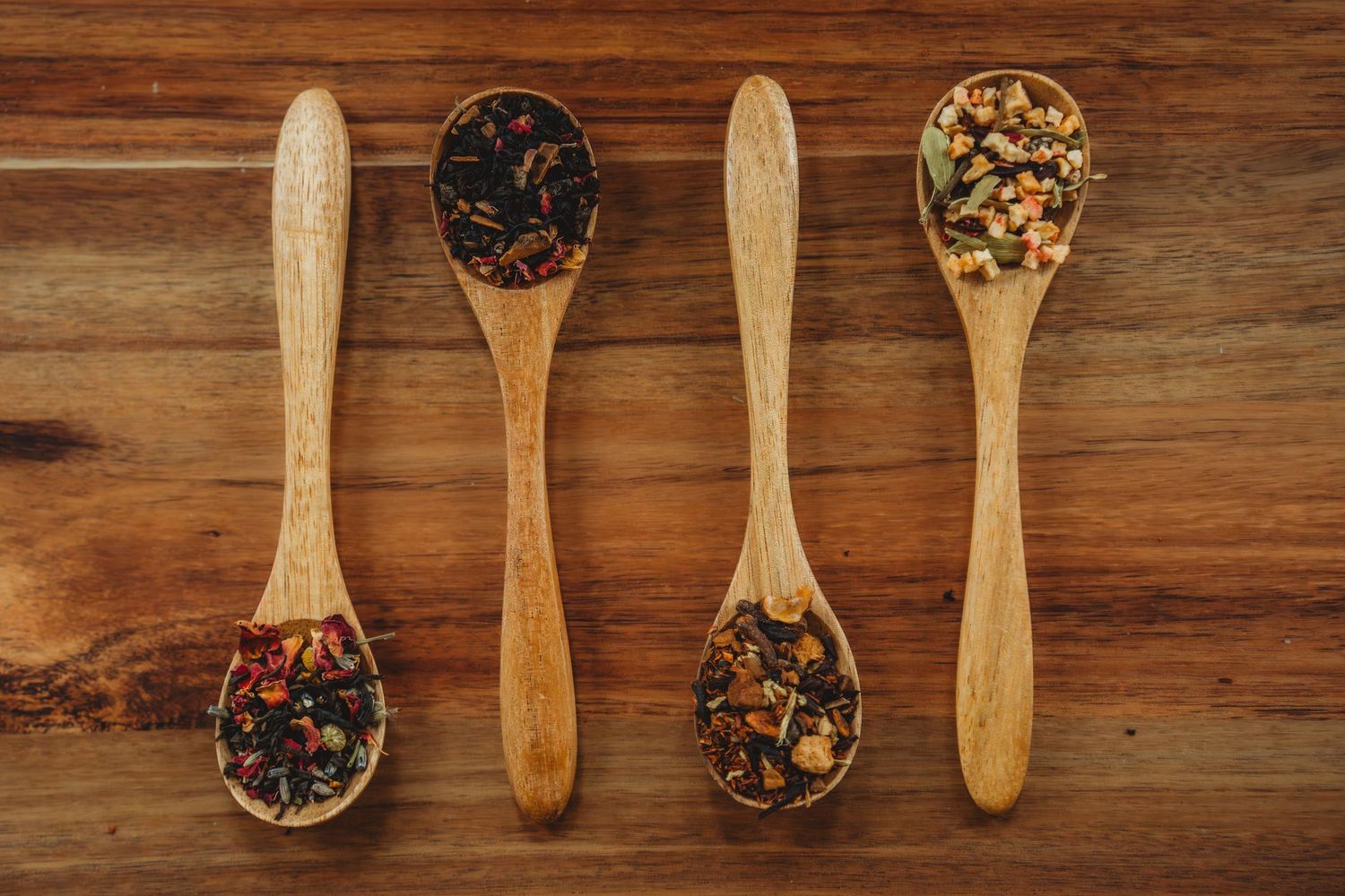 Four wooden spoons with different varieties of loose leaf herbal tea displayed on rustic wood surface.