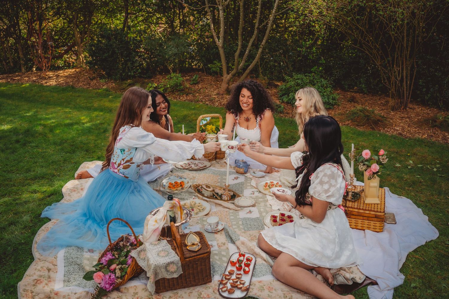Group of women in white dresses enjoying a picnic on a blanket in a lush garden.
