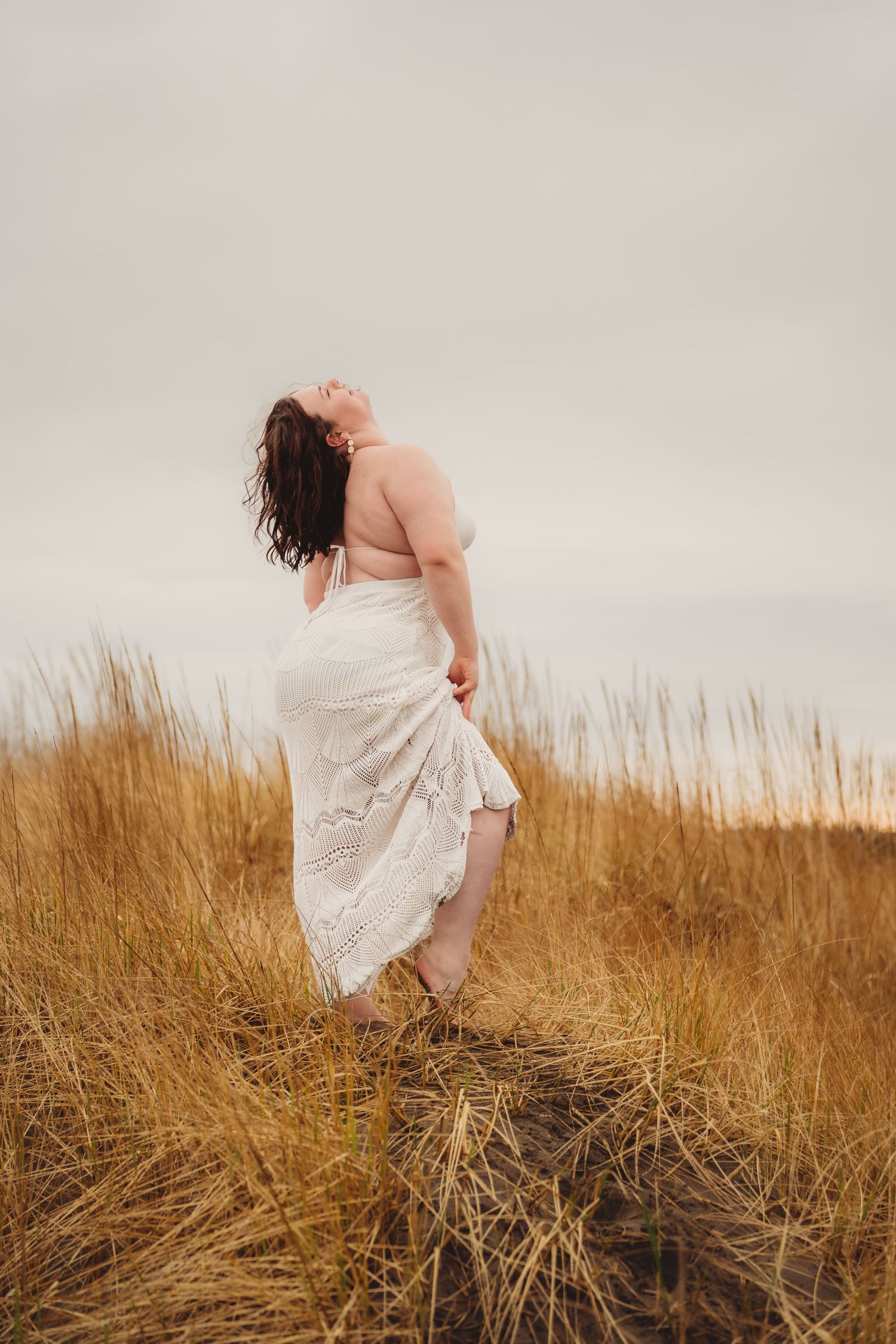 Woman in white lace dress standing in golden grass field, head tilted back, hair flowing in breeze.