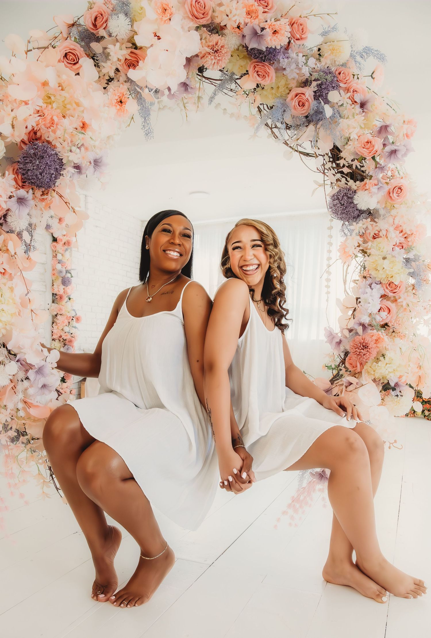 Two brides in white dresses sit back-to-back smiling before a pastel floral arch with pink, lavender, and peach blooms.