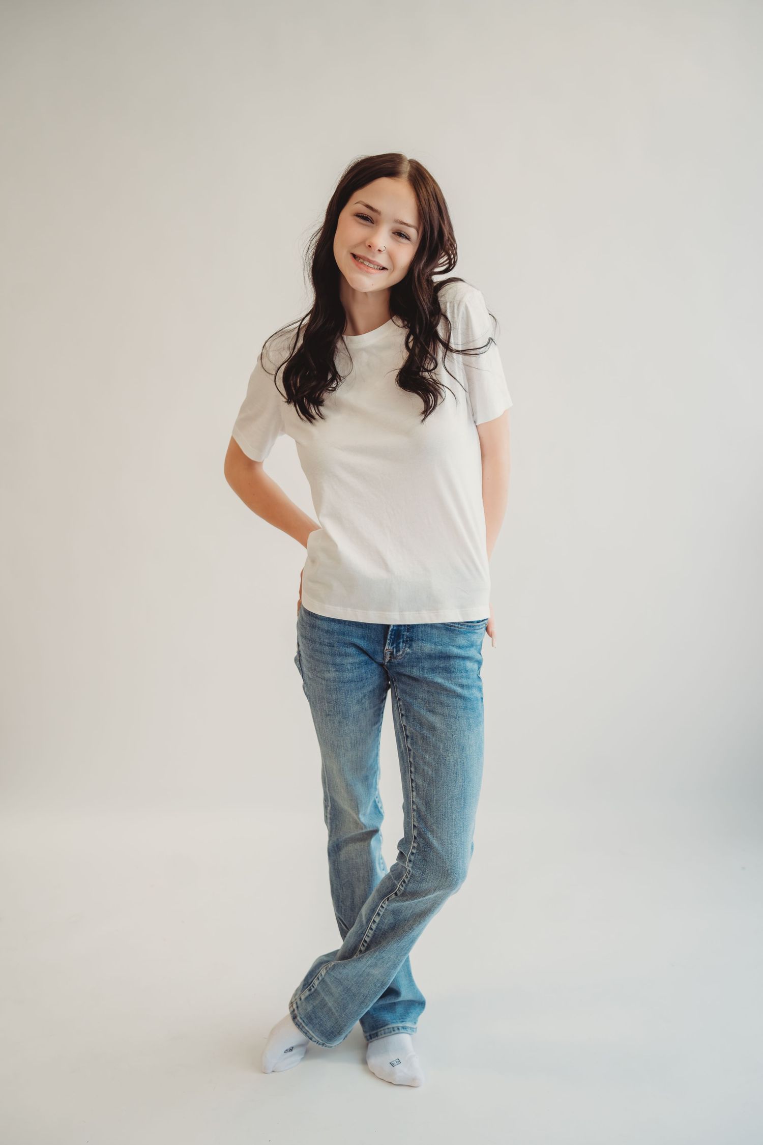 Young woman in white t-shirt and blue jeans smiling, standing against a light gray background.