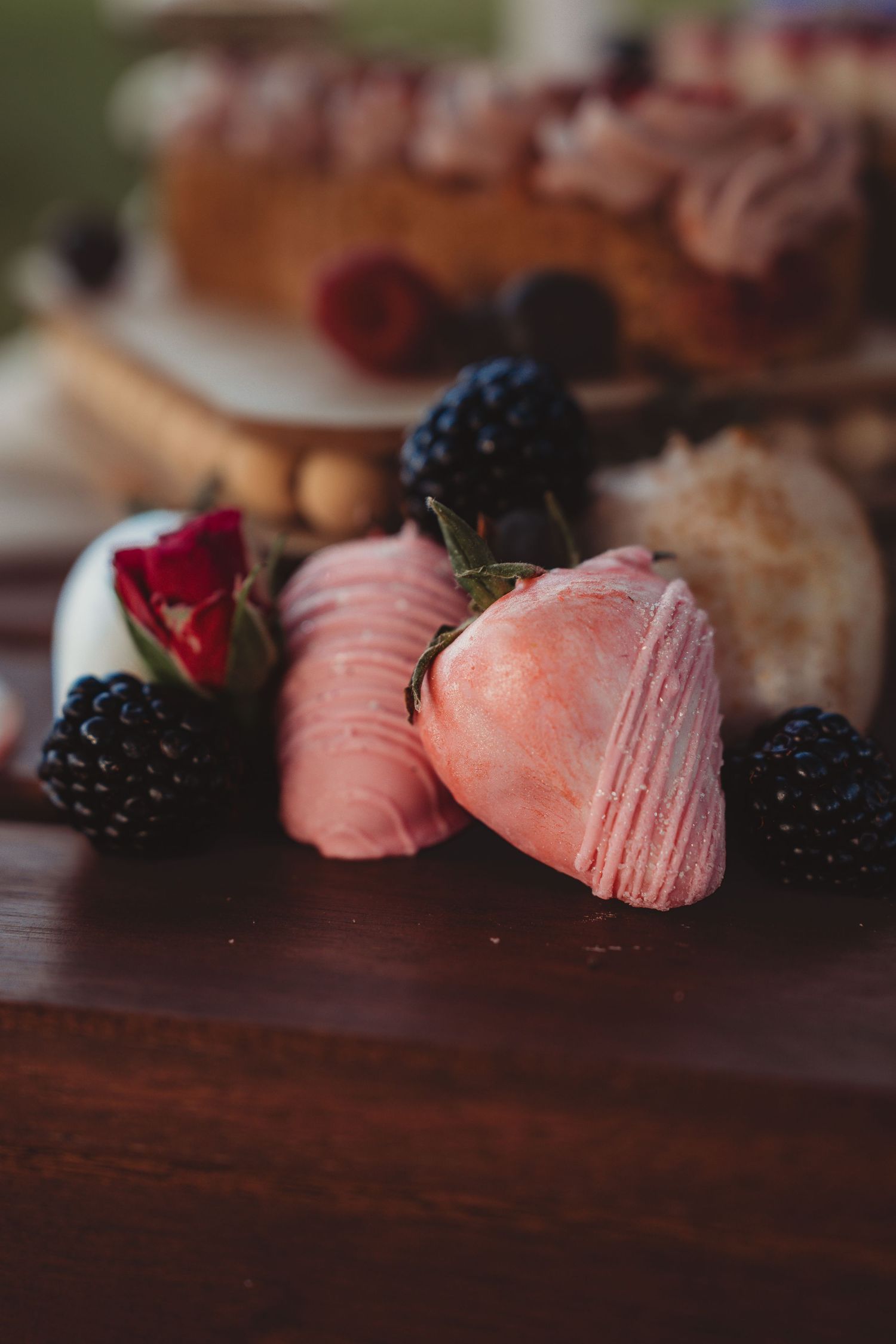 Pink chocolate-dipped strawberries with blackberries and a rose on a wooden board, with a berry cake in the background.