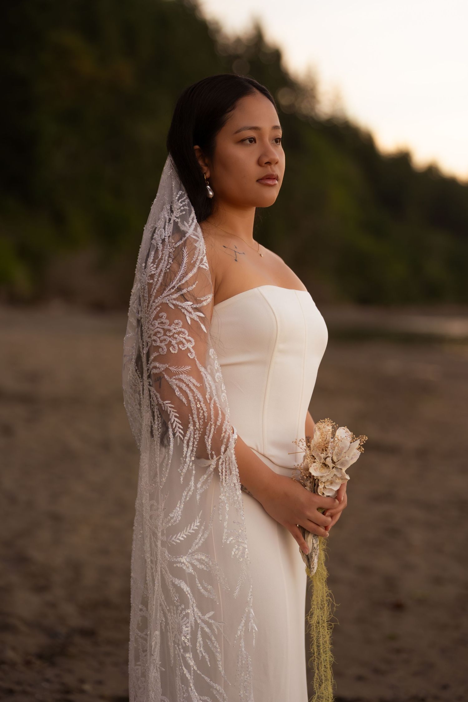Bride in white strapless gown and lace veil holds bouquet on beach at sunset.
