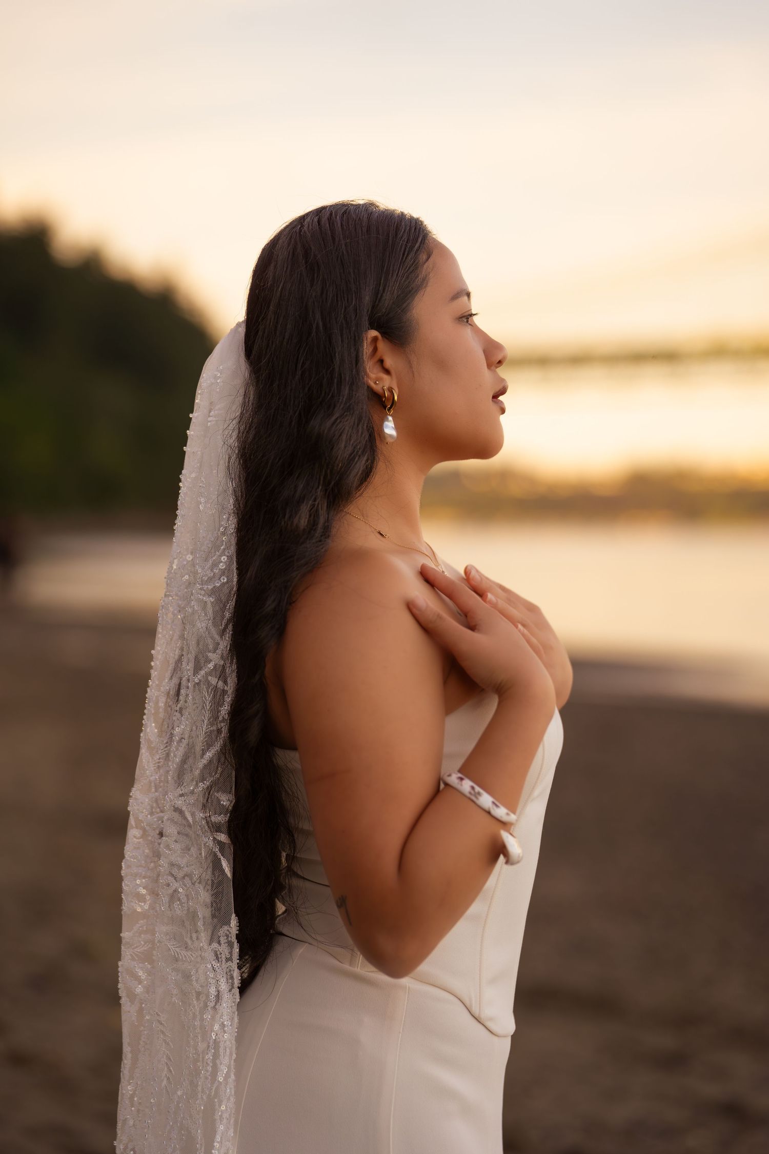 Bride in white strapless gown and lace veil poses at sunset beach, hands crossed over chest, long dark hair.