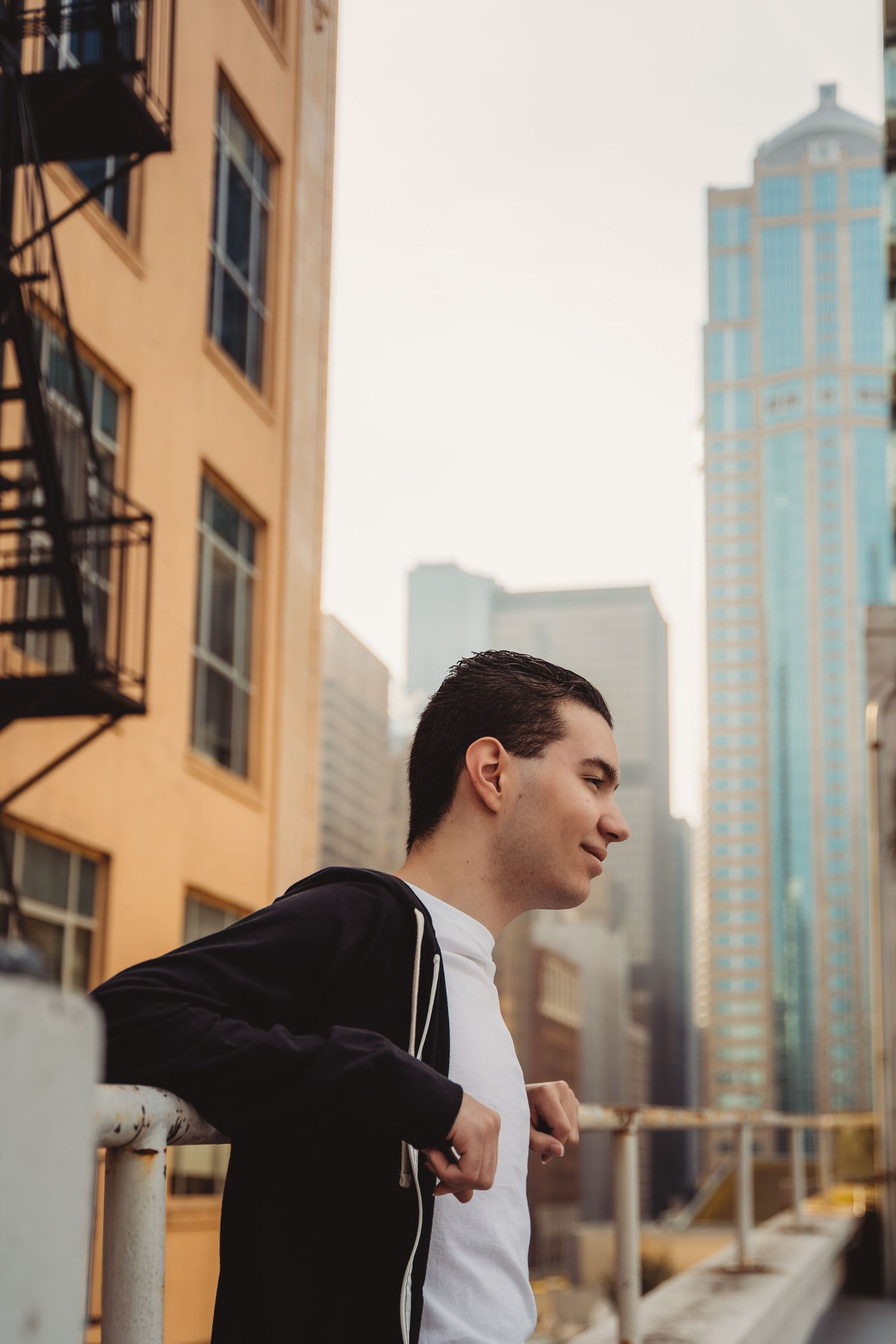 Young man in black jacket leaning on railing with city skyscrapers in background on urban rooftop.