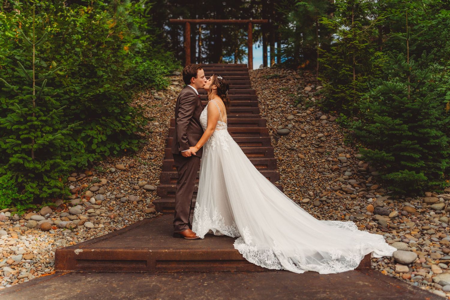 Bride and groom sharing a kiss on wooden steps surrounded by lush greenery, bride wearing elegant white gown with long train.