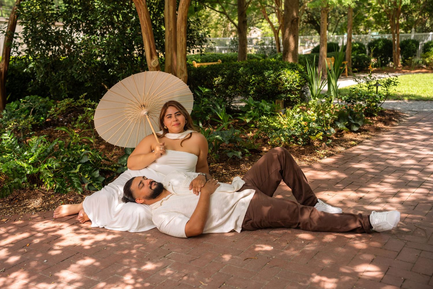 A couple relaxes on a brick path in a lush garden, woman holding a parasol umbrella, wearing white and brown outfits.