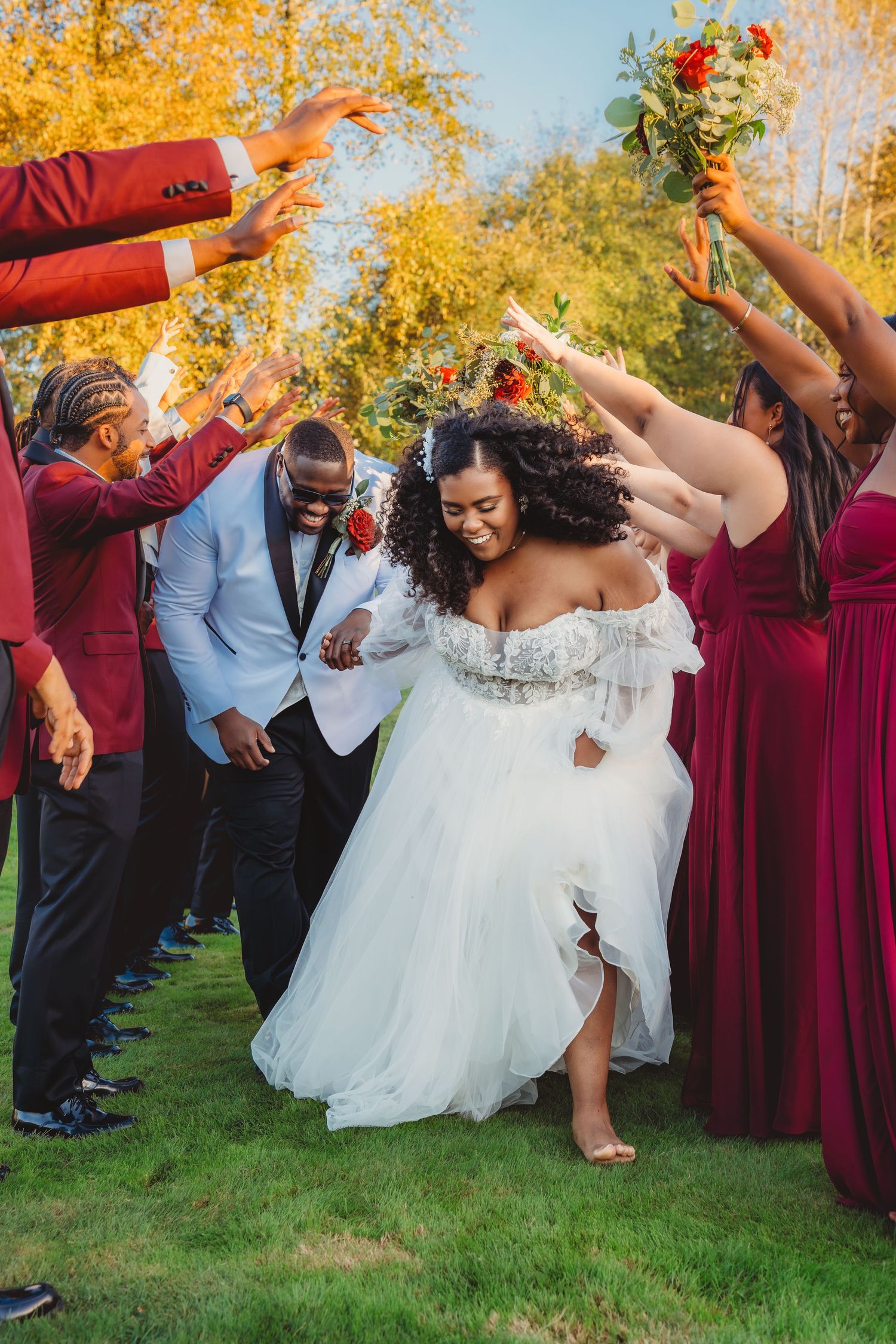 Joyful Black bride and groom walk through a celebratory arch of hands with wedding party in burgundy attire outdoors.