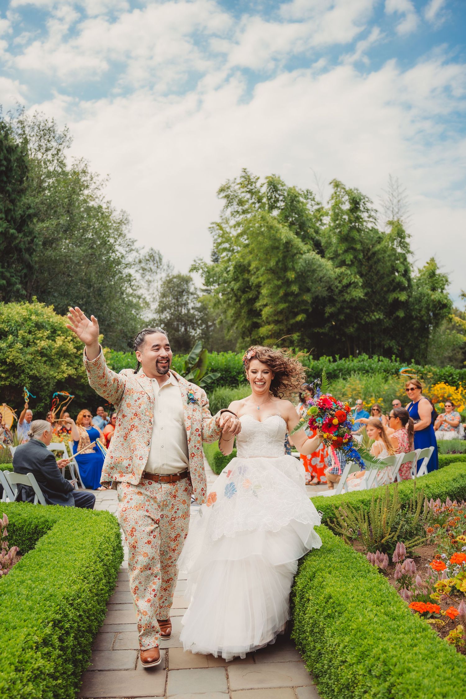 Joyful newlyweds walk down garden aisle, groom in floral suit waving, bride in white tiered gown holding colorful bouquet.