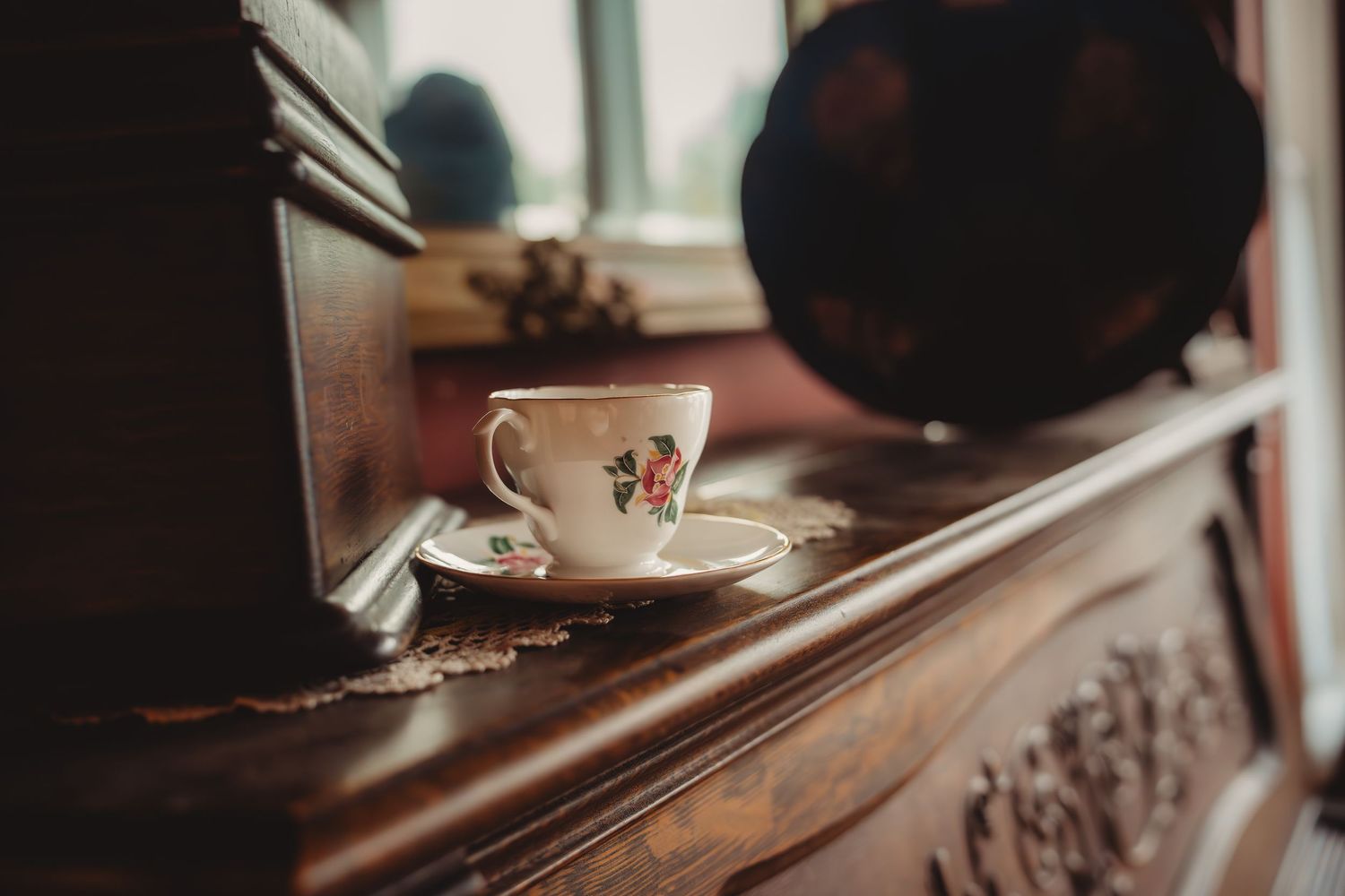Vintage floral teacup and saucer on ornate dark wood dresser near window with soft natural light.