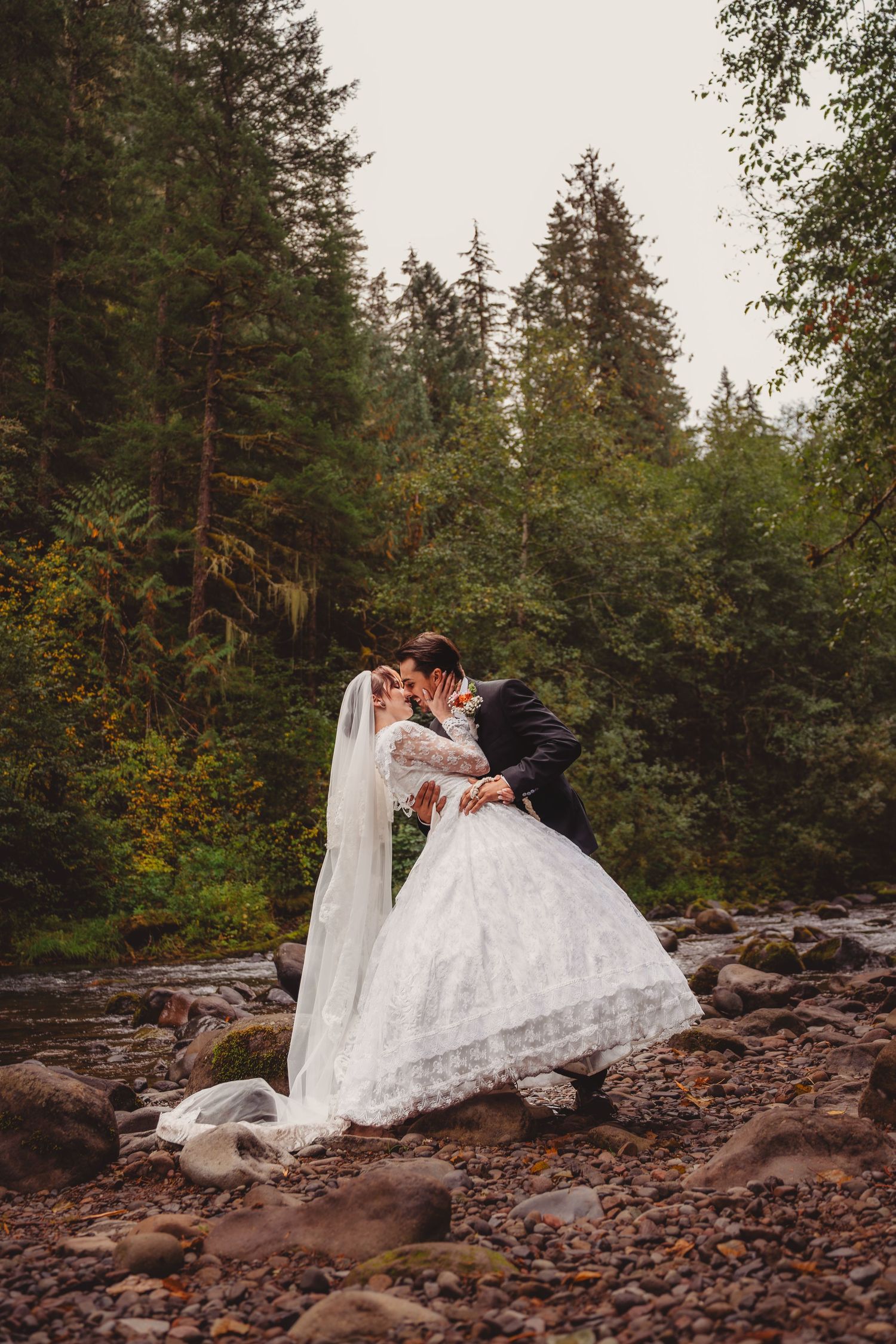 Bride and groom share a romantic kiss on rocky riverbank surrounded by lush evergreen forest in outdoor wedding photo. mount hood Oregon