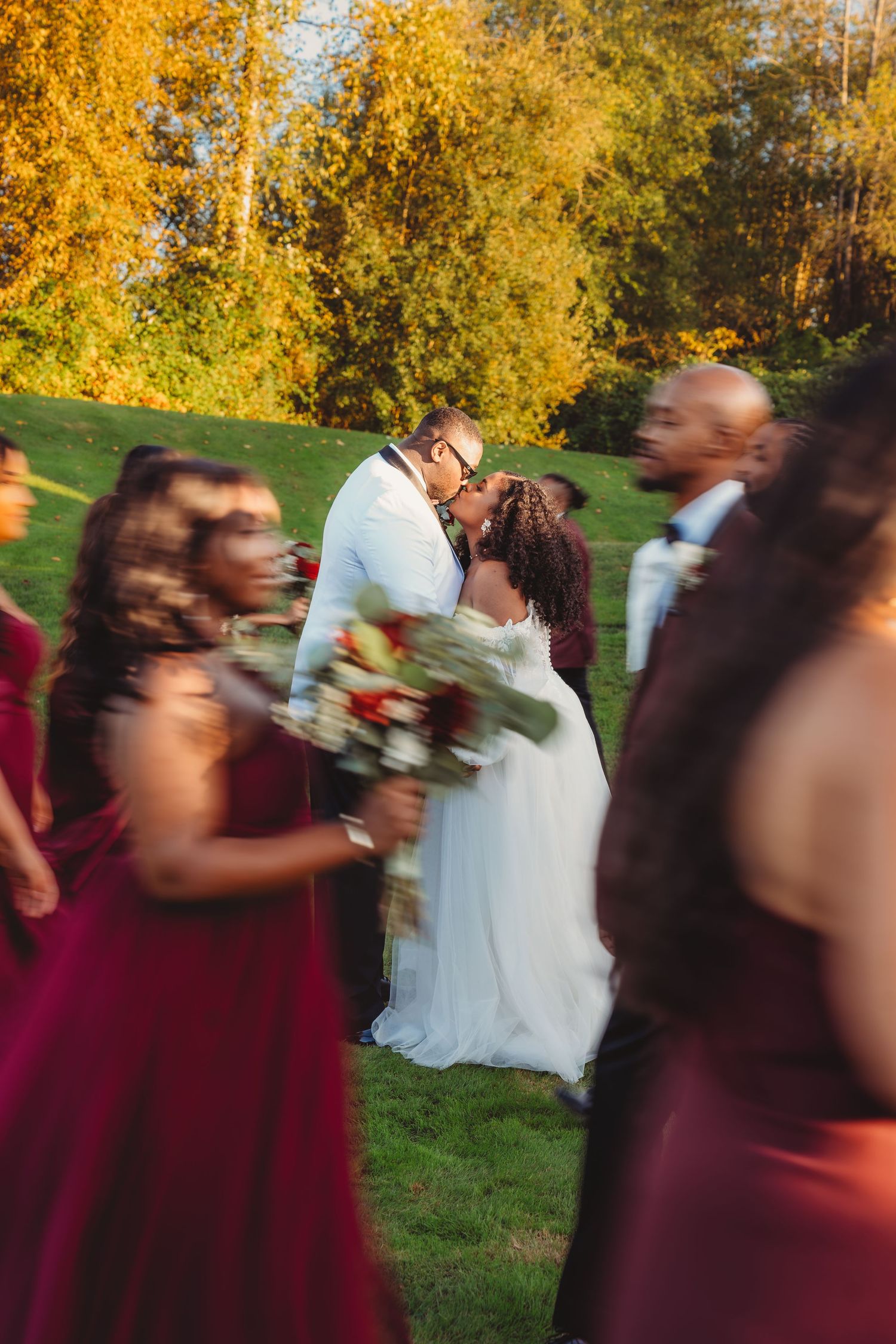 Bride and groom kissing surrounded by wedding party in burgundy dresses outdoors. South Carolina golf course