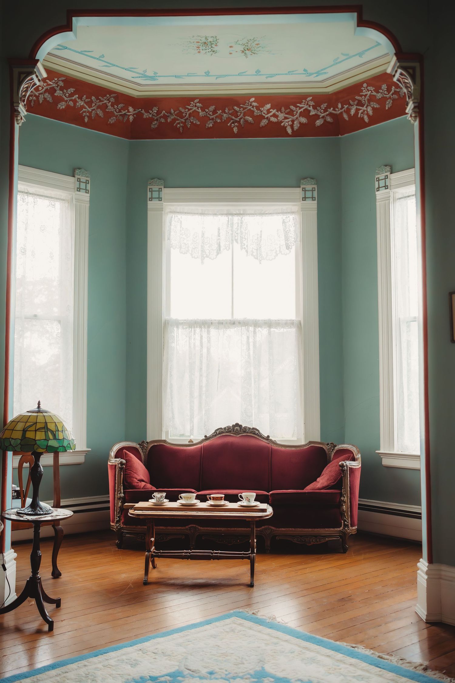 Elegant Victorian parlor with red velvet sofa, ornate ceiling molding, bay windows, and vintage Tiffany lamp.