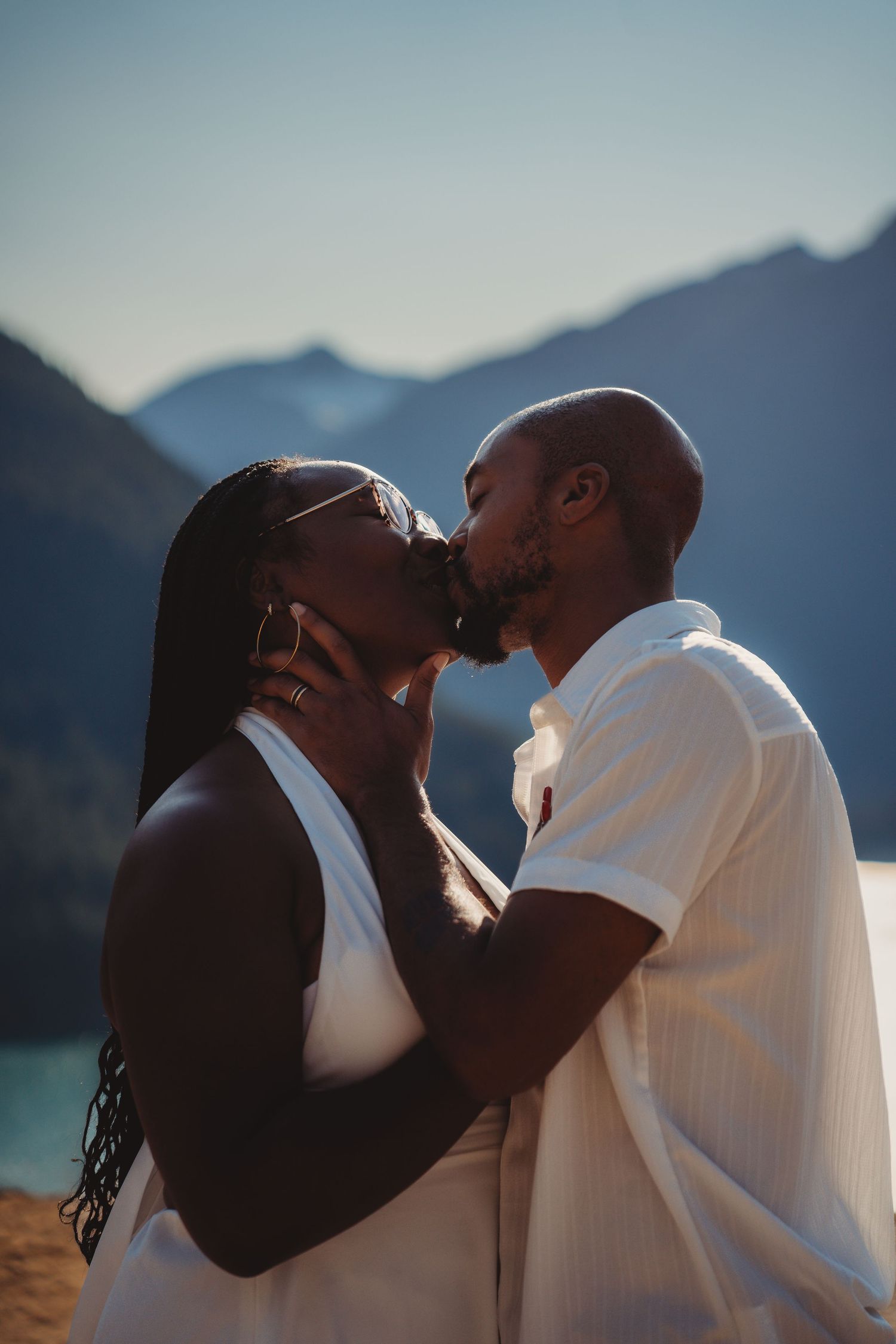 A couple sharing a romantic kiss outdoors with mountains and a lake in the background.