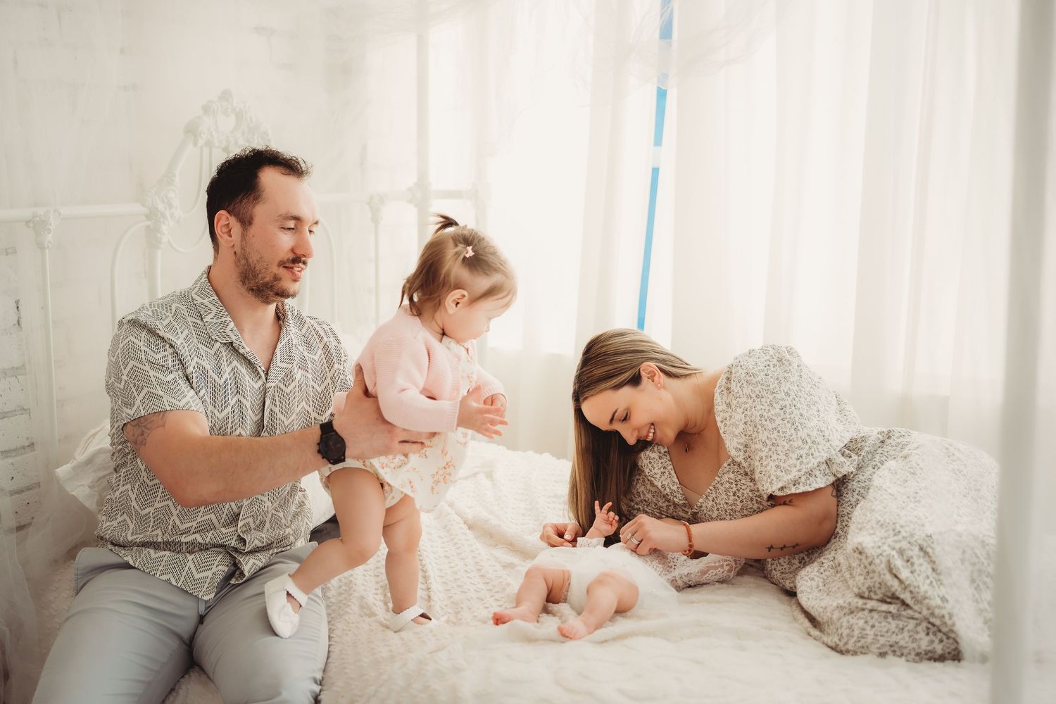 Happy family with toddler and newborn baby relaxing together on white bed in bright cozy bedroom.