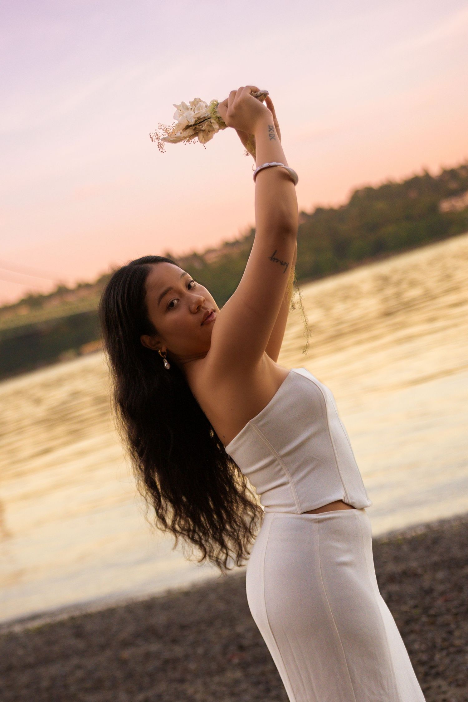 Young woman in white outfit holding flowers above head at sunset by a lake.