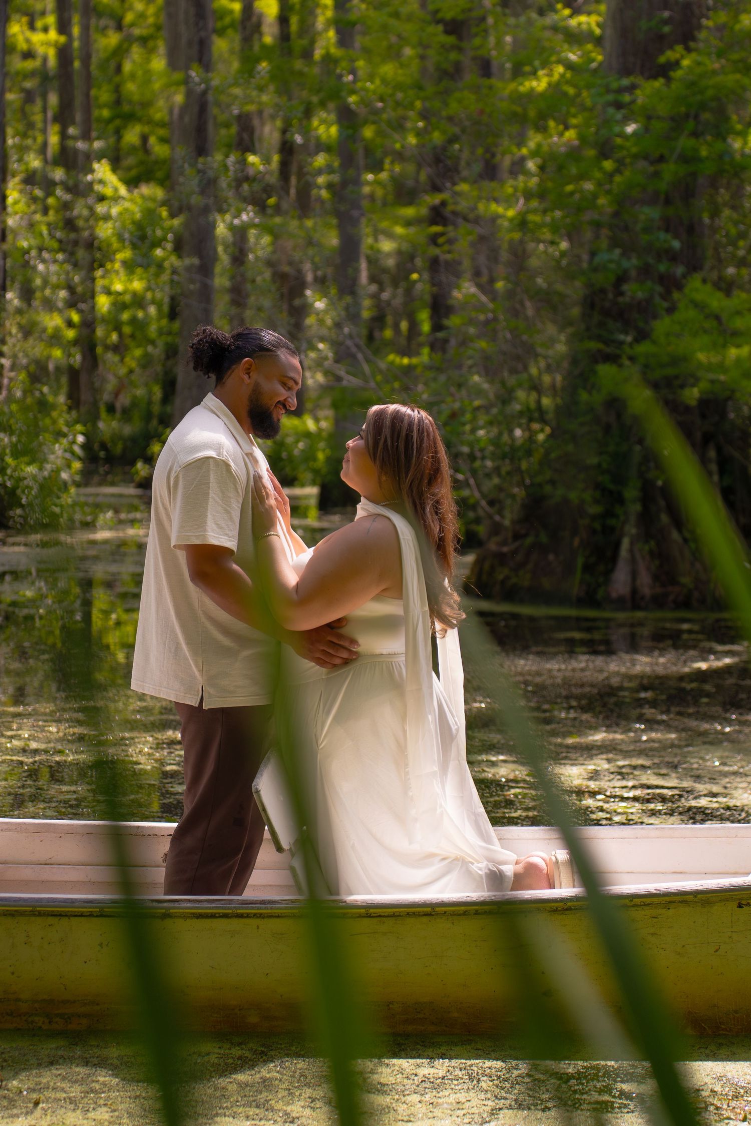 A couple shares a romantic moment on a boat in a lush green swamp, the woman in white dress, man in casual attire.