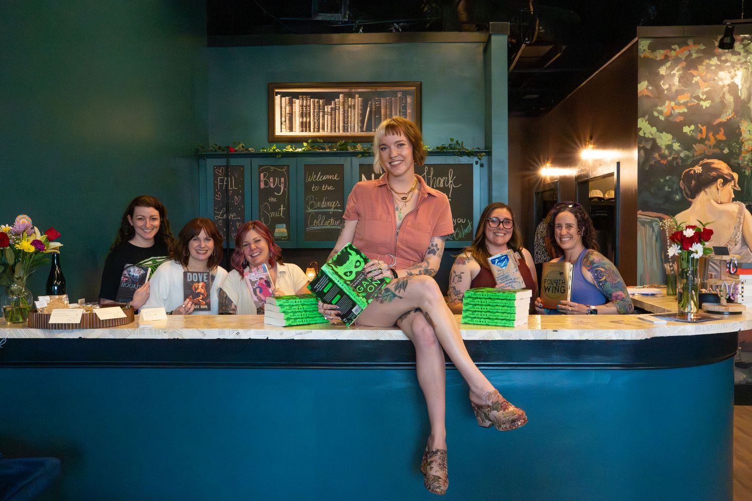 Group of smiling women holding green books gathered around teal bookstore counter display.