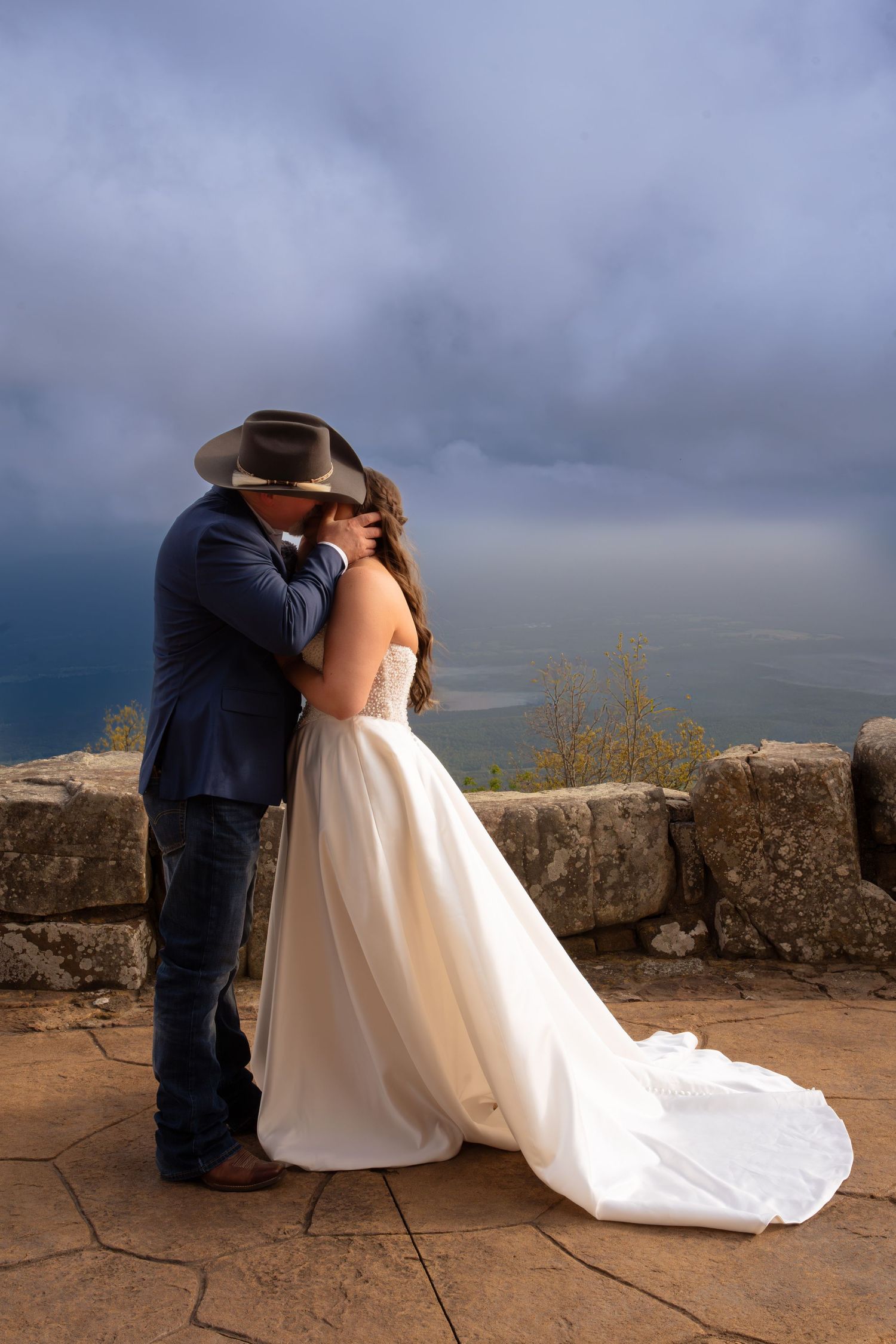 Bride and groom kissing at mountain overlook, groom in cowboy hat and jeans, bride in white gown, stormy sky backdrop.