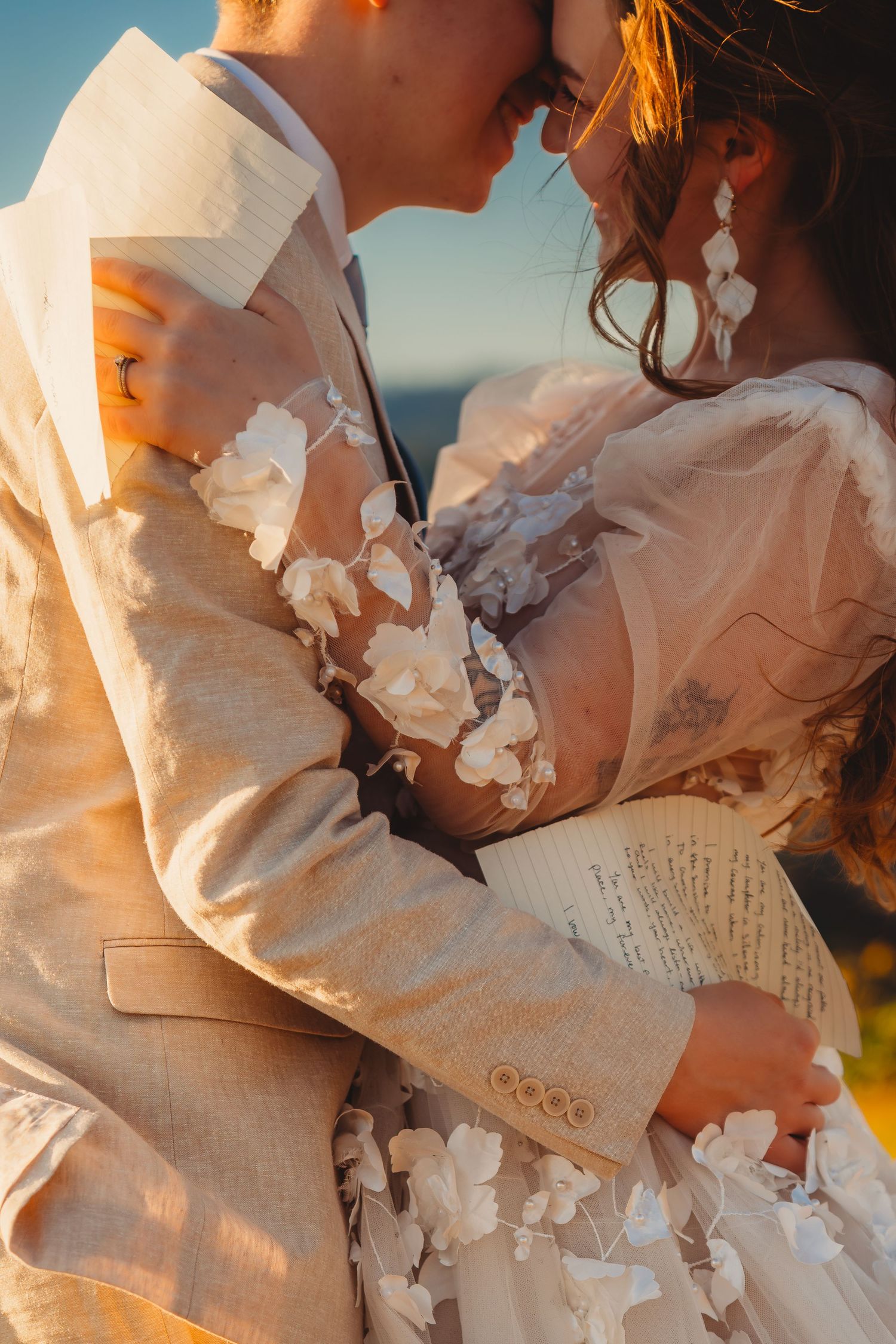 Couple in wedding attire embracing closely at golden hour, bride in floral applique gown, groom in beige suit.
