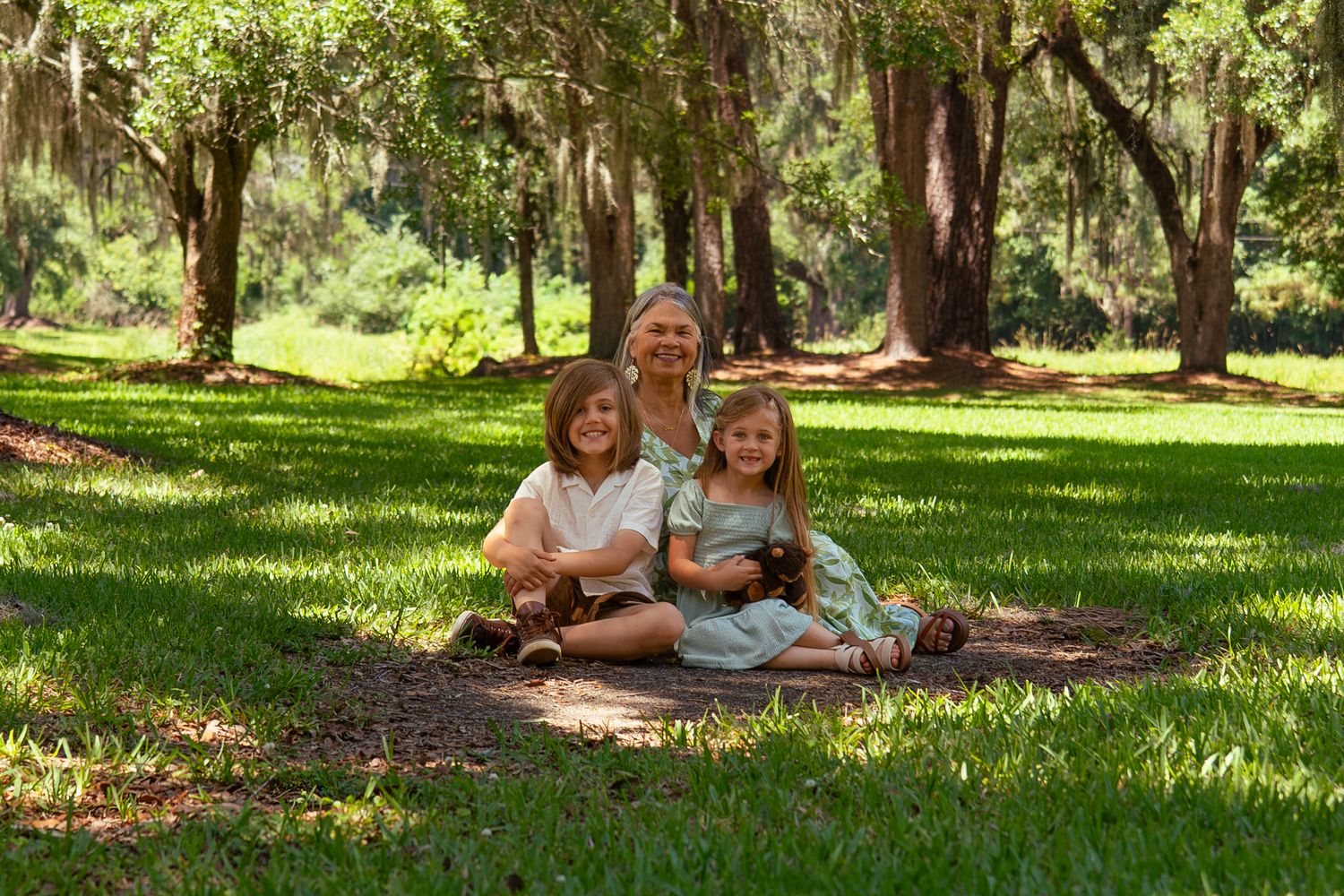 A parent sitting on grass with two young children smiling in a lush green park setting.