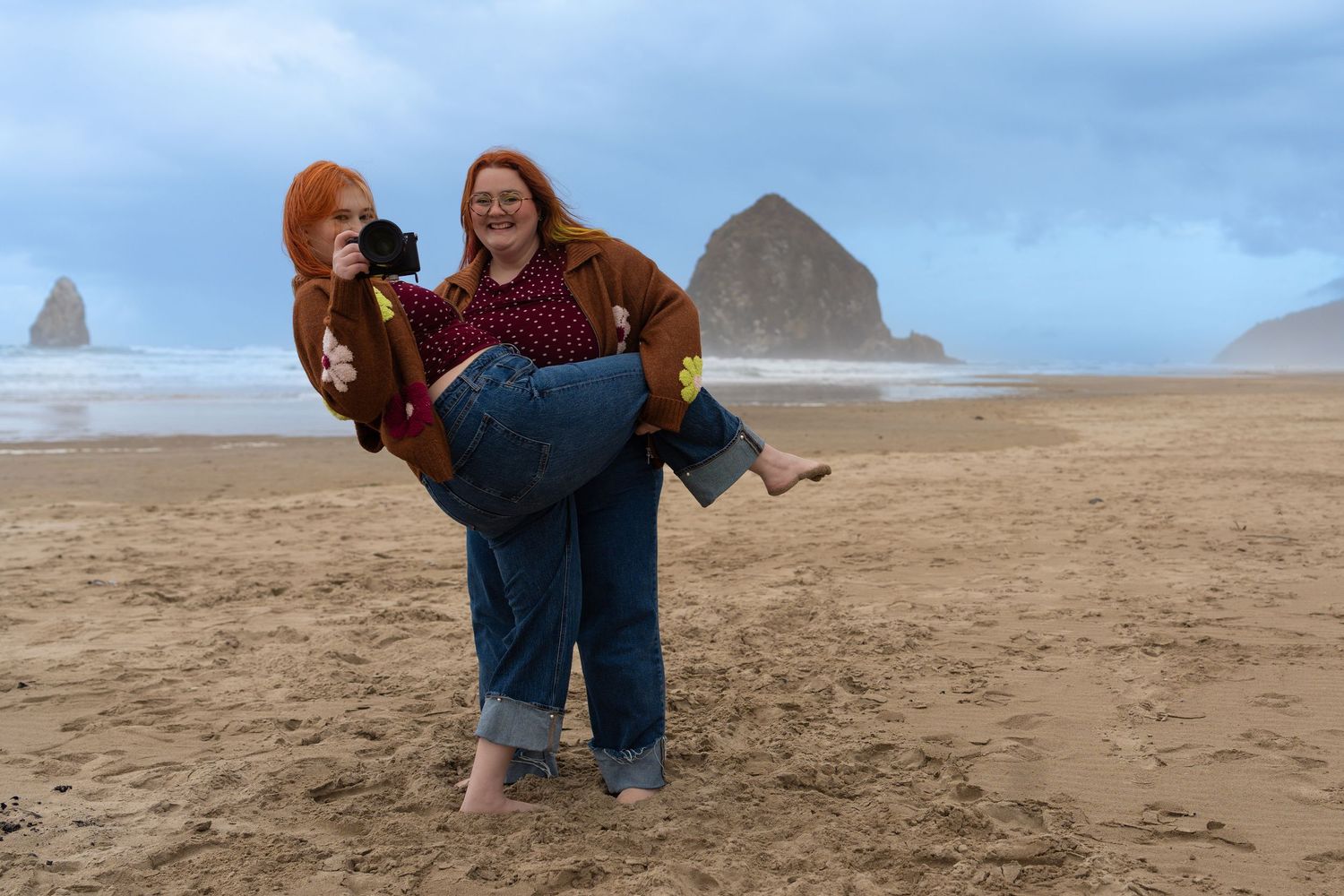 Two women laughing on Cannon Beach, Oregon, with Haystack Rock in the background on a cloudy day.
