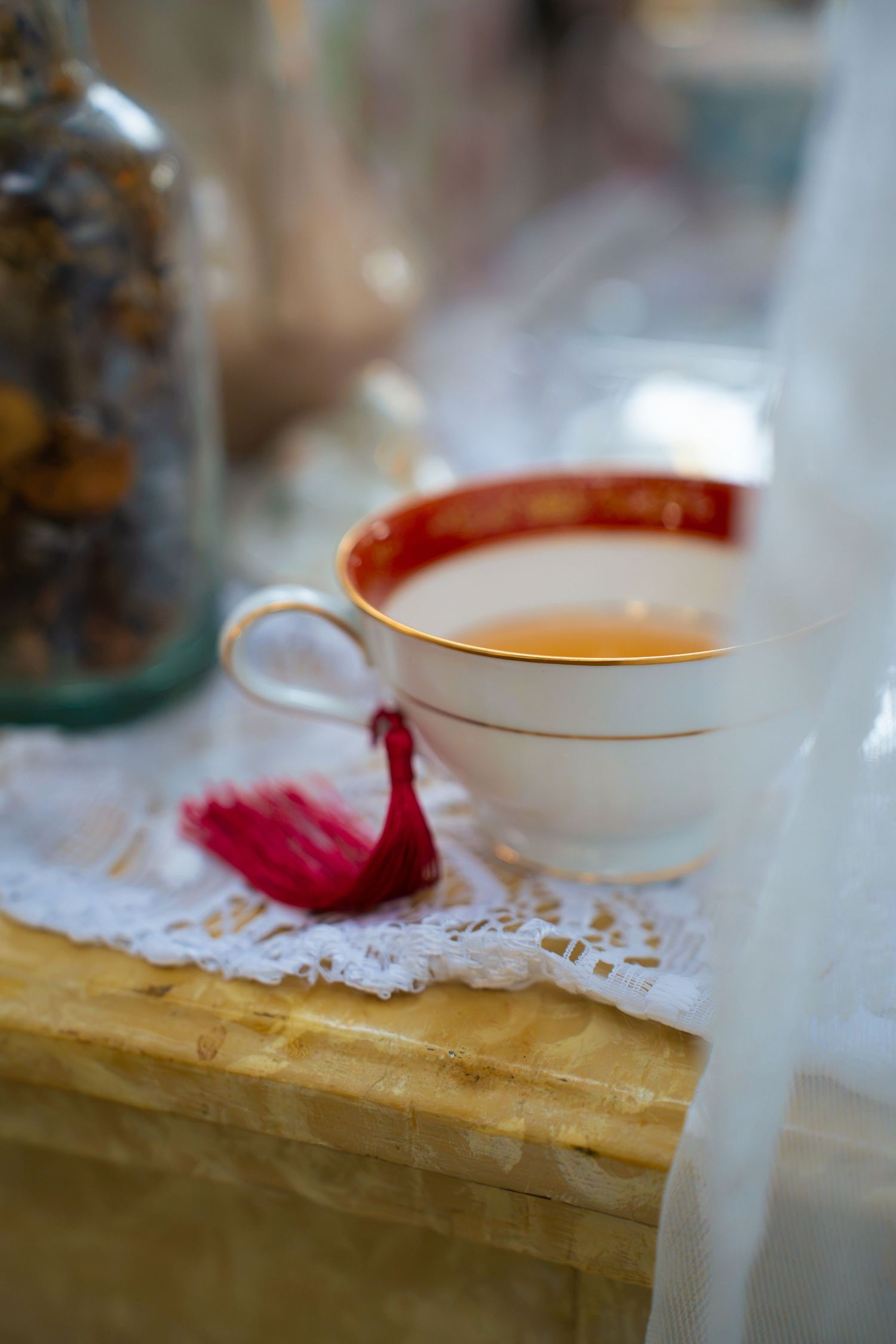 White porcelain teacup with red and gold rim filled with tea, resting on lace doily beside a red tassel and a jar of dried tea leaves.