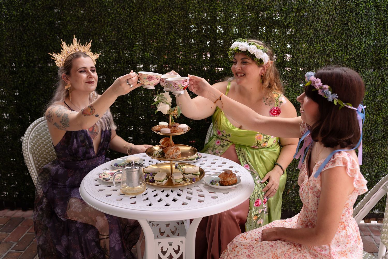 Three women in floral crowns and fairy-tale gowns toast at an outdoor tea party with tiered pastry stand.