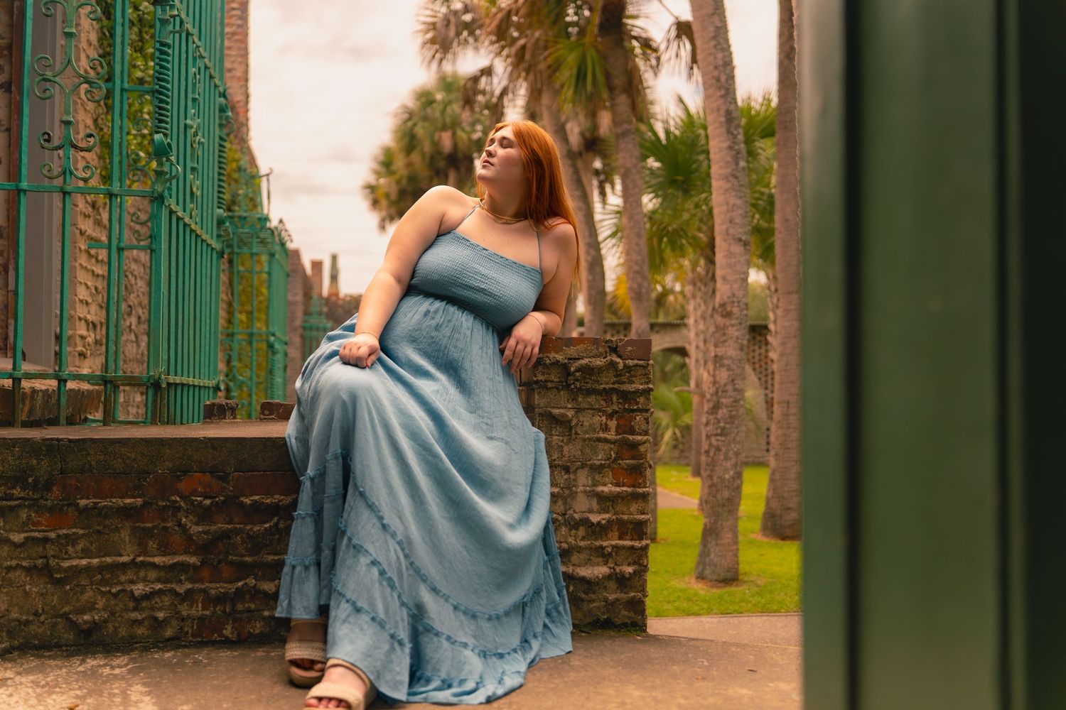 A woman in a flowing blue maxi dress sits on a stone wall surrounded by palm trees and iron gates.