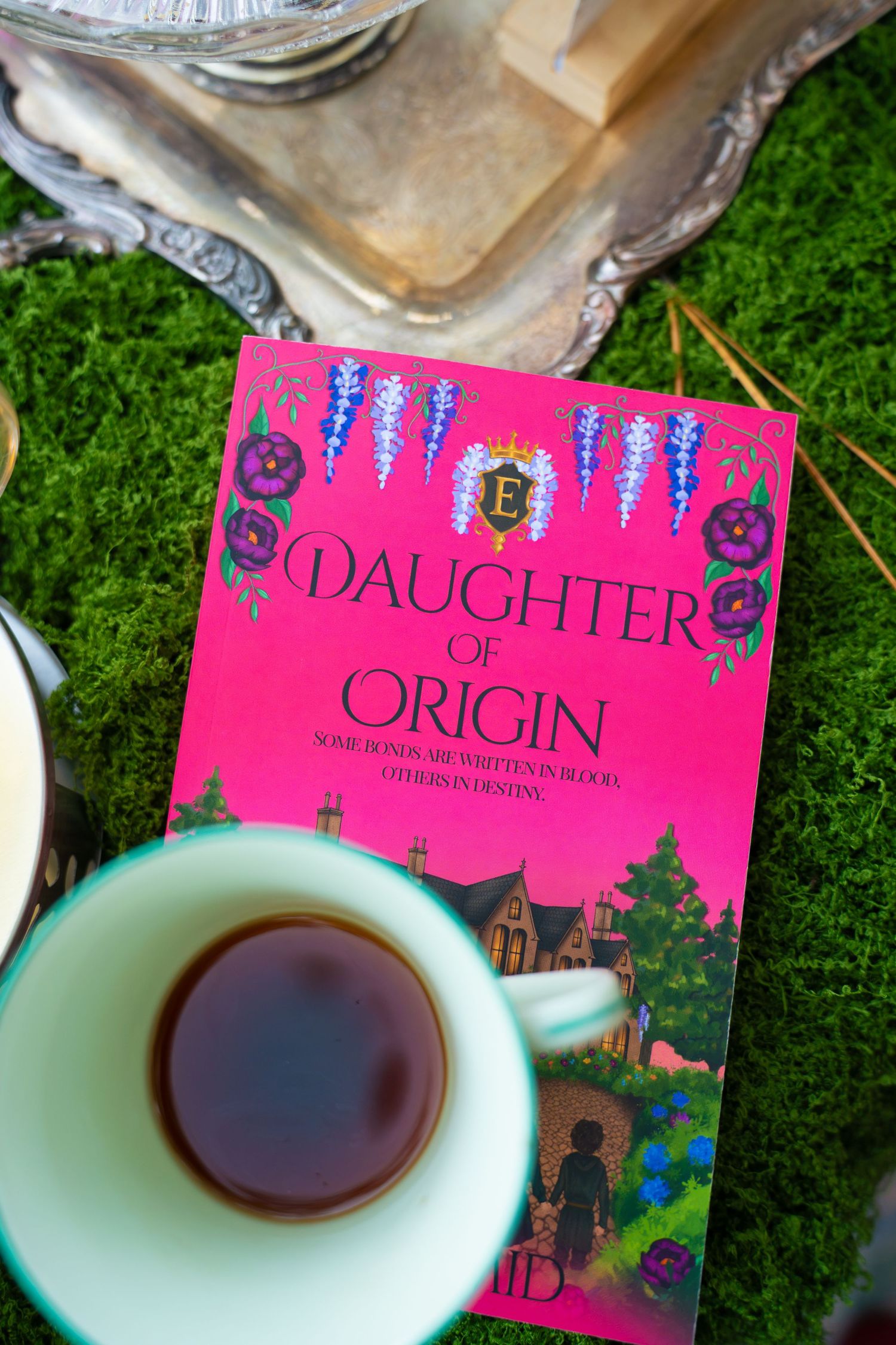 Pink book Daughter of Origin with purple flowers rests on green moss beside a cup of tea and an ornate silver tray.