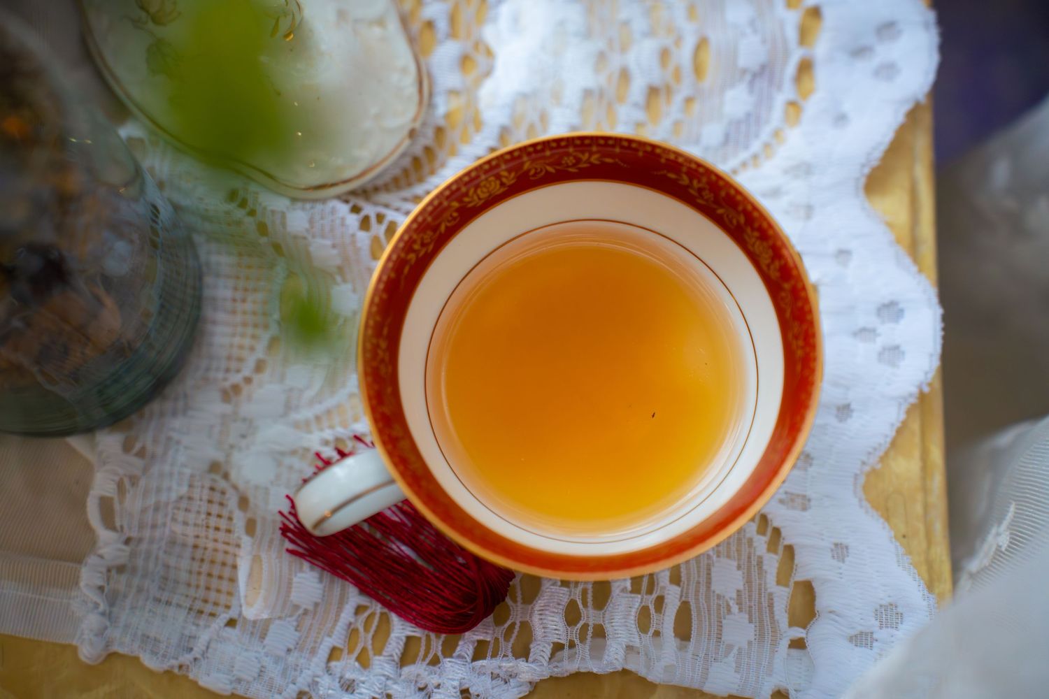 Overhead view of a golden herbal tea in an orange-rimmed porcelain cup on white lace doily, beside glass jars and green leaves.
