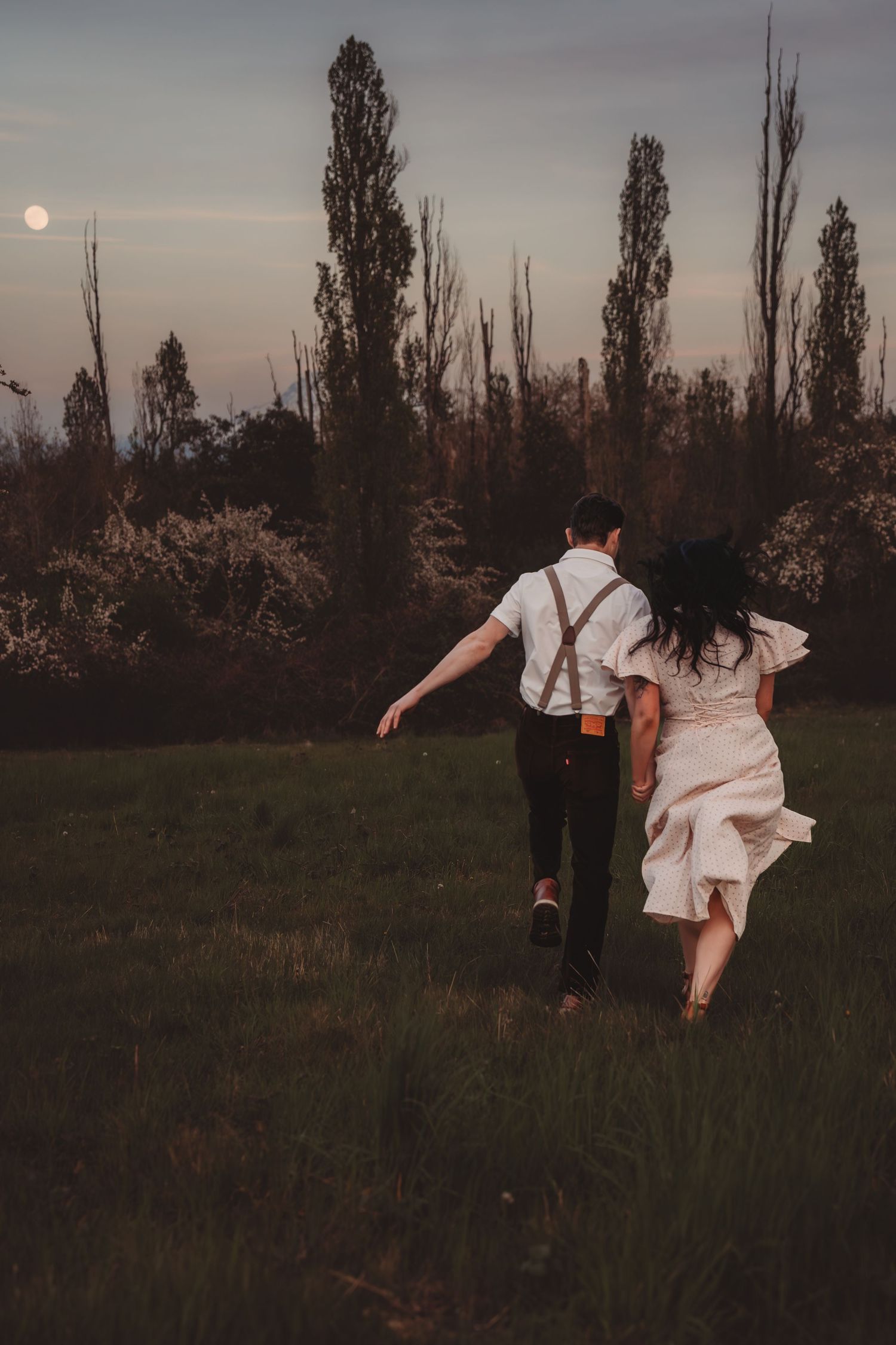 Couple in wedding attire running through a moonlit field at dusk, surrounded by trees and blooming shrubs.