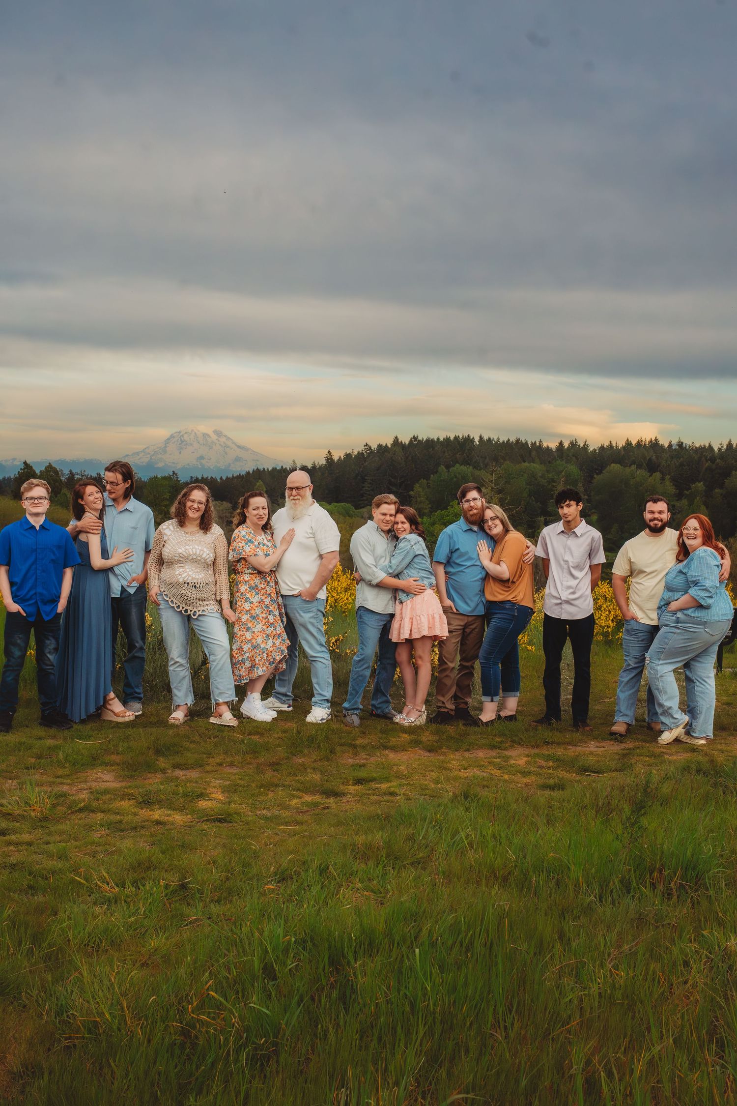 Large family group posing outdoors in a green field with Mount Rainier and forest backdrop under cloudy skies.
