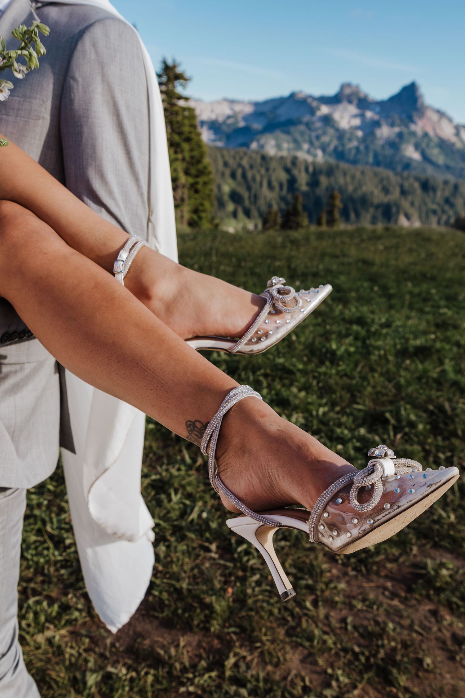 Close-up of bride wearing elegant embellished heels with ankle strap, mountain landscape visible in background.