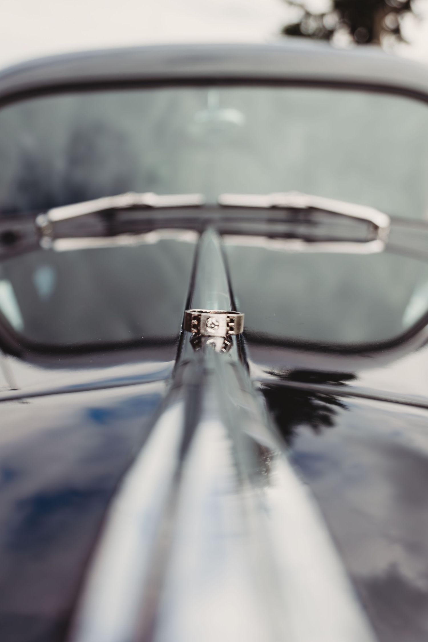 A diamond engagement ring balanced on the hood ornament of a vintage classic car with chrome details.