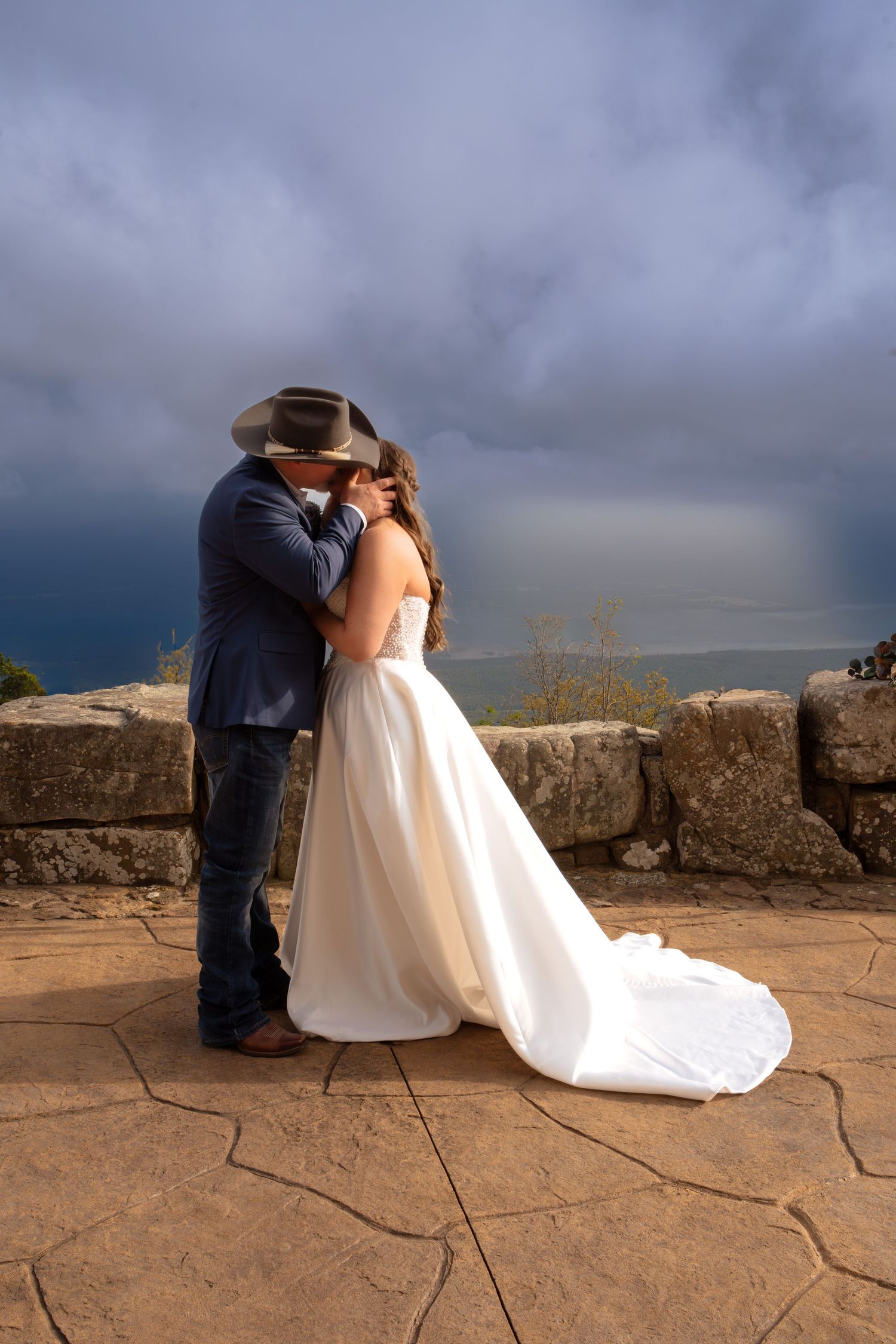 Bride and groom kissing outdoors in stormy weather, wearing cowboy hat and white wedding gown with train.