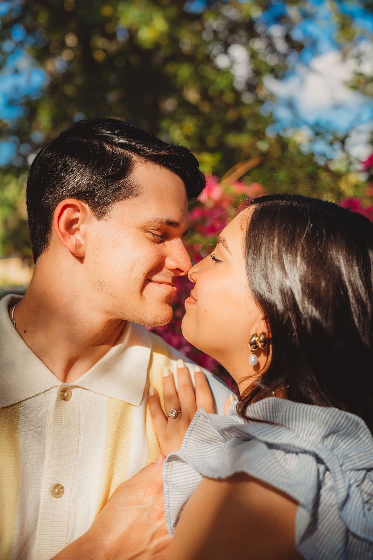 Romantic couple sharing a tender almost-kiss outdoors, surrounded by lush greenery and pink flowers in warm sunlight.