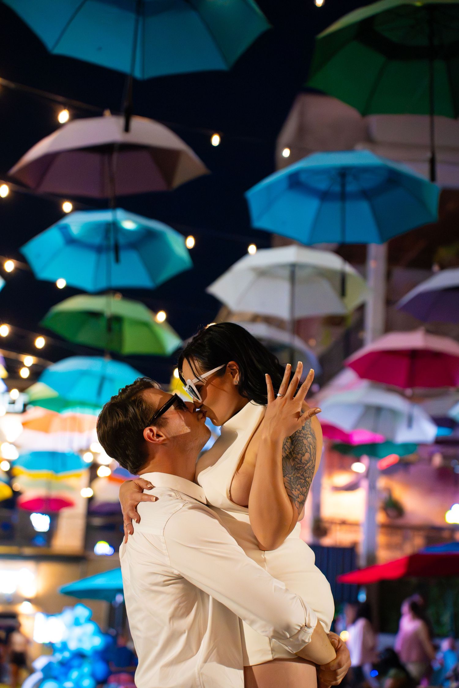 Couple in sunglasses embracing and kissing under colorful hanging umbrellas and string lights at a lively evening outdoor venue.