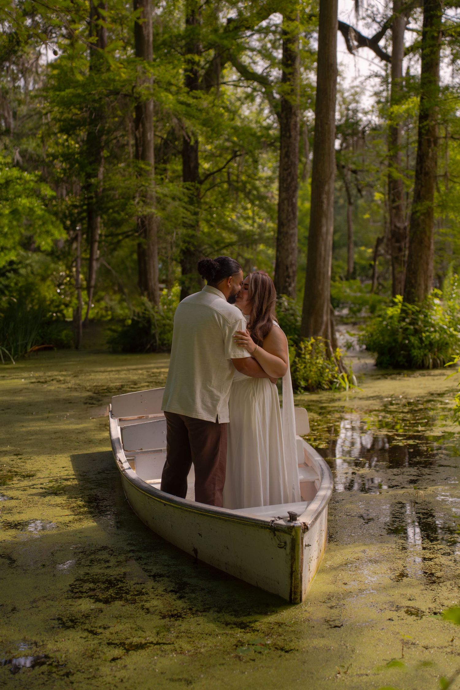 A couple shares a romantic kiss while standing in a white canoe surrounded by a lush, green cypress swamp.