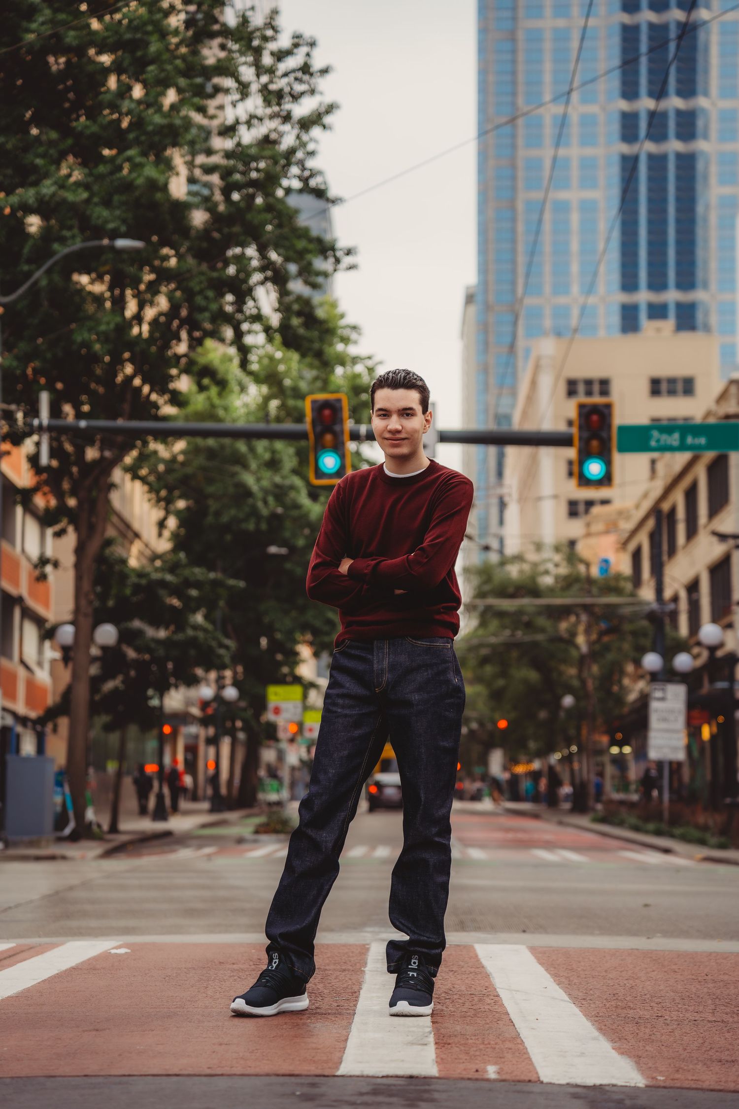 Young man in red sweater and jeans stands confidently on urban crosswalk with city buildings and green traffic lights behind him.