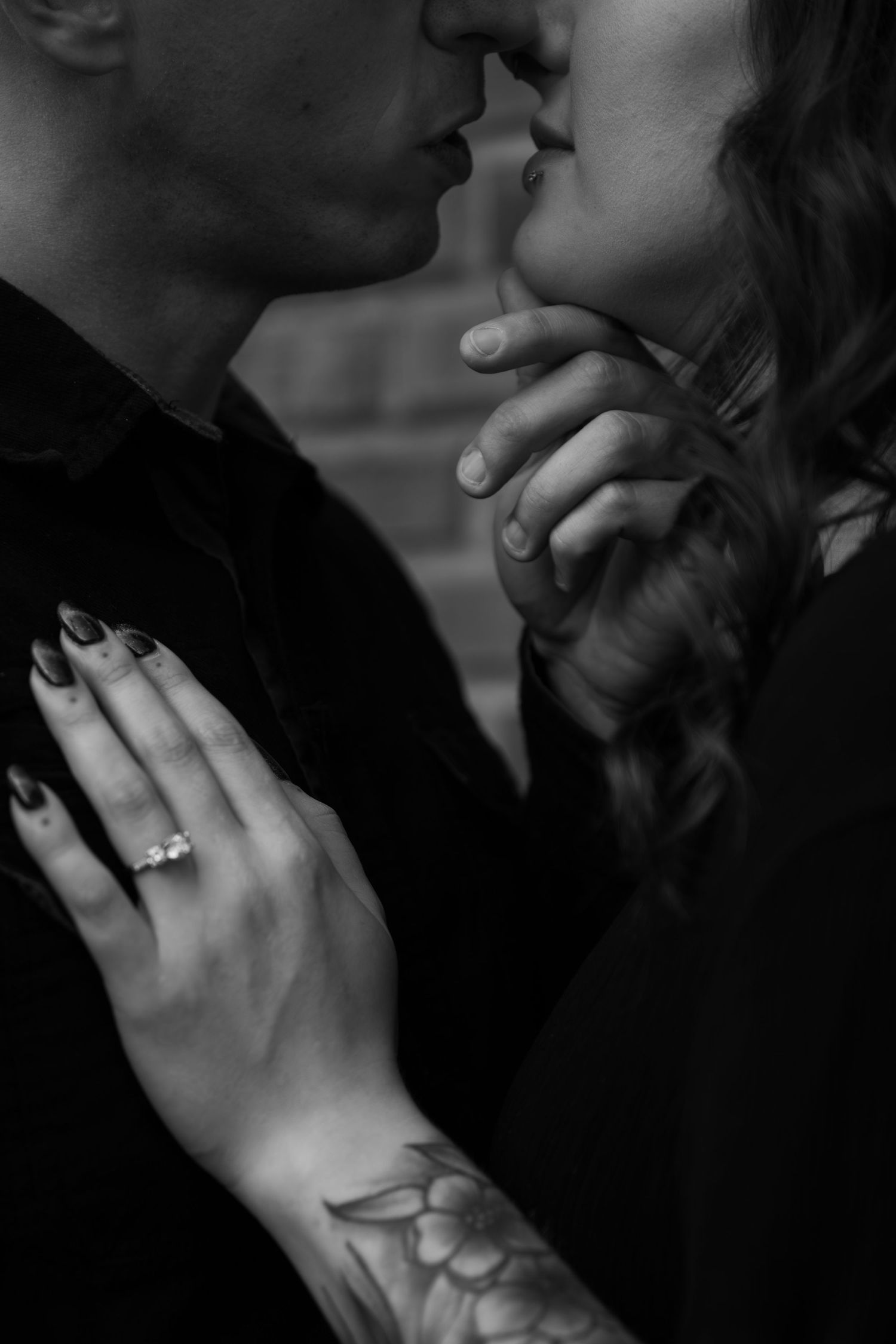 Black and white photo of a couple kissing, woman showing diamond engagement ring with tattooed arm visible.