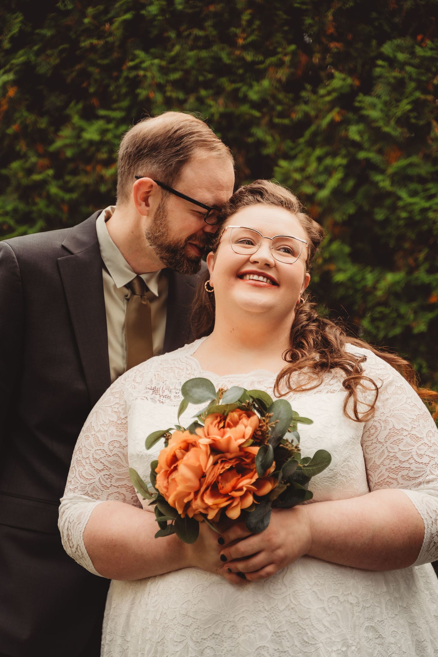 A groom in a dark suit whispers to his smiling bride in white lace holding orange roses bouquet outdoors.