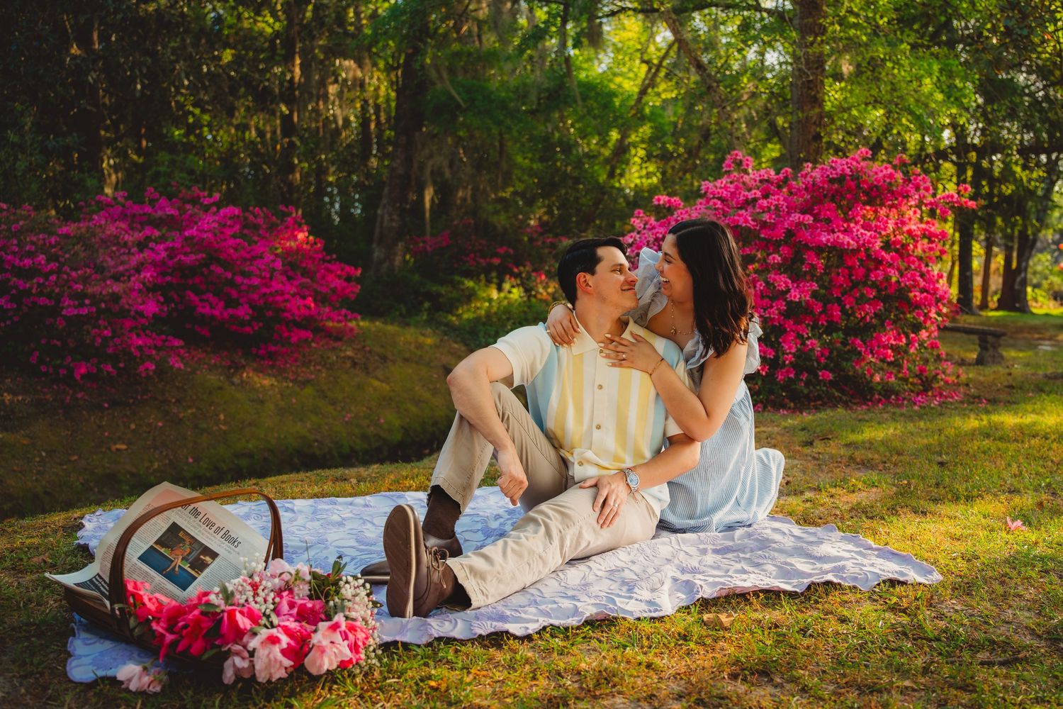 Couple sharing a romantic kiss on a picnic blanket surrounded by blooming pink azaleas in a lush garden.