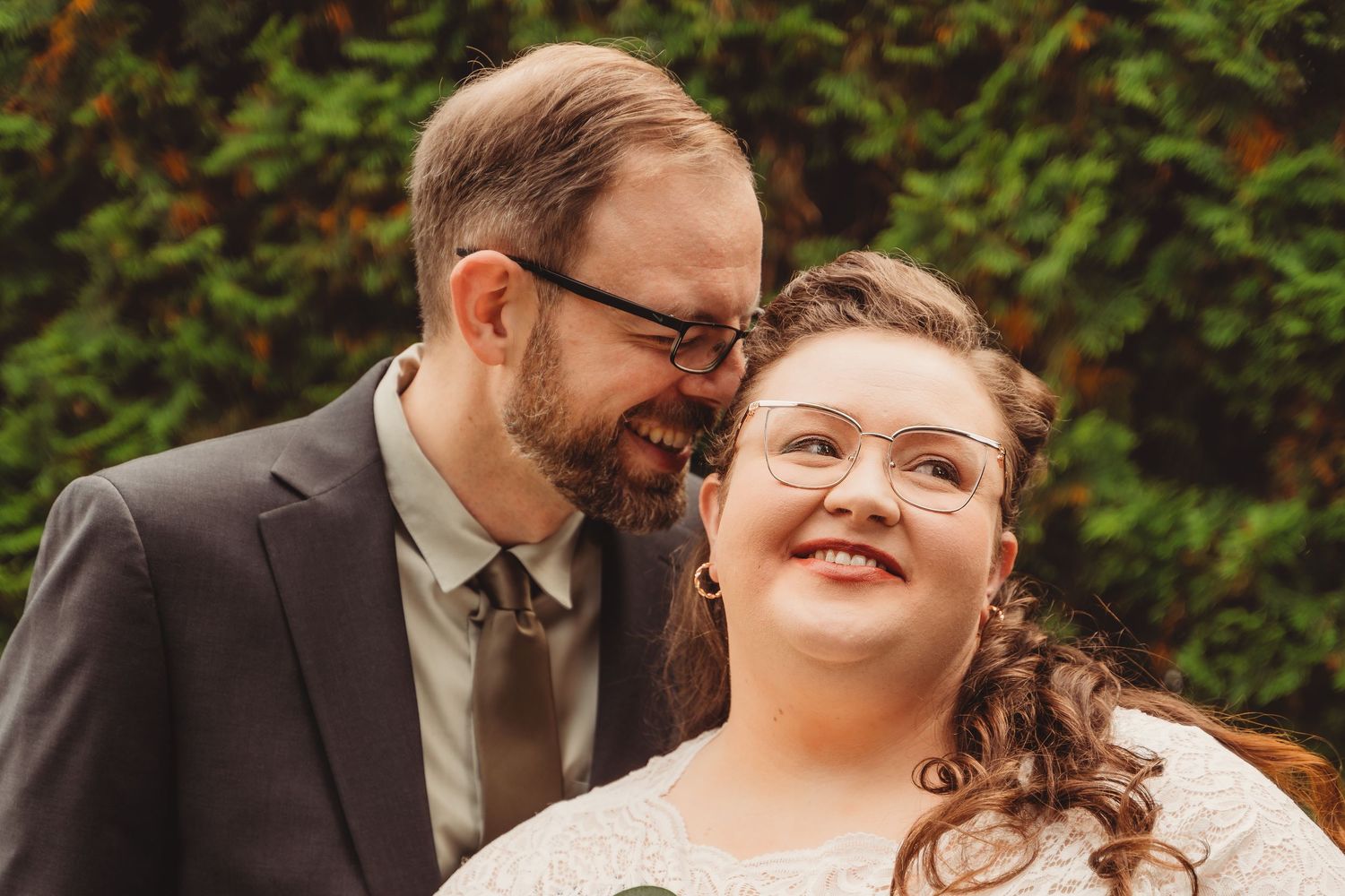 Happy couple smiling closely together outdoors, groom in gray suit whispering to smiling bride wearing glasses. Oregon